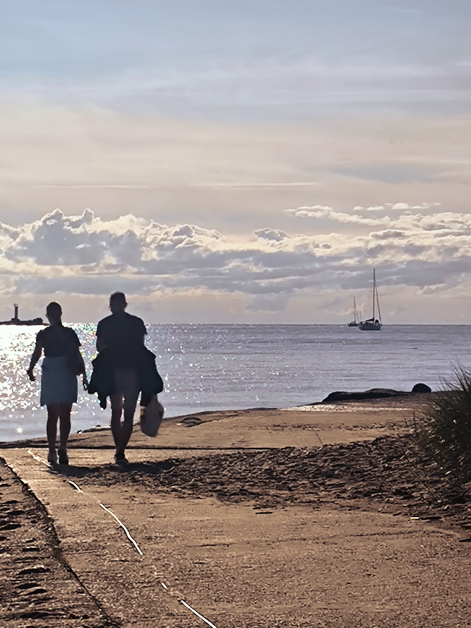 Two figures, silhouetted against a sparkling ocean, walk along a paved path bordering a sandy beach. The figure on the left, appearing to be a woman, wears a light-colored skirt and a darker top. She carries no visible objects. The figure on the right, appearing to be a man, wears dark clothing and carries a light-colored bag in his right hand and a dark jacket over his left shoulder. Both figures are walking away from the camera.
The ocean stretches out before them, reflecting the light of a hazy sky filled with scattered clouds. Two sailboats are visible on the horizon. To the left, a lighthouse stands on a distant landmass. The path is made of concrete with visible seams, and sand is present on either side. Vegetation, appearing as tall, dry grass, is visible on the far right of the frame.
The scene is consistent with the location of Daugava River mouth, near the Baltic Sea, in Riga, Latvia. The lighthouse in the distance resembles the one at the end of the Juras Street promenade in Vecaki. The overall lighting suggests it is late afternoon or early evening.