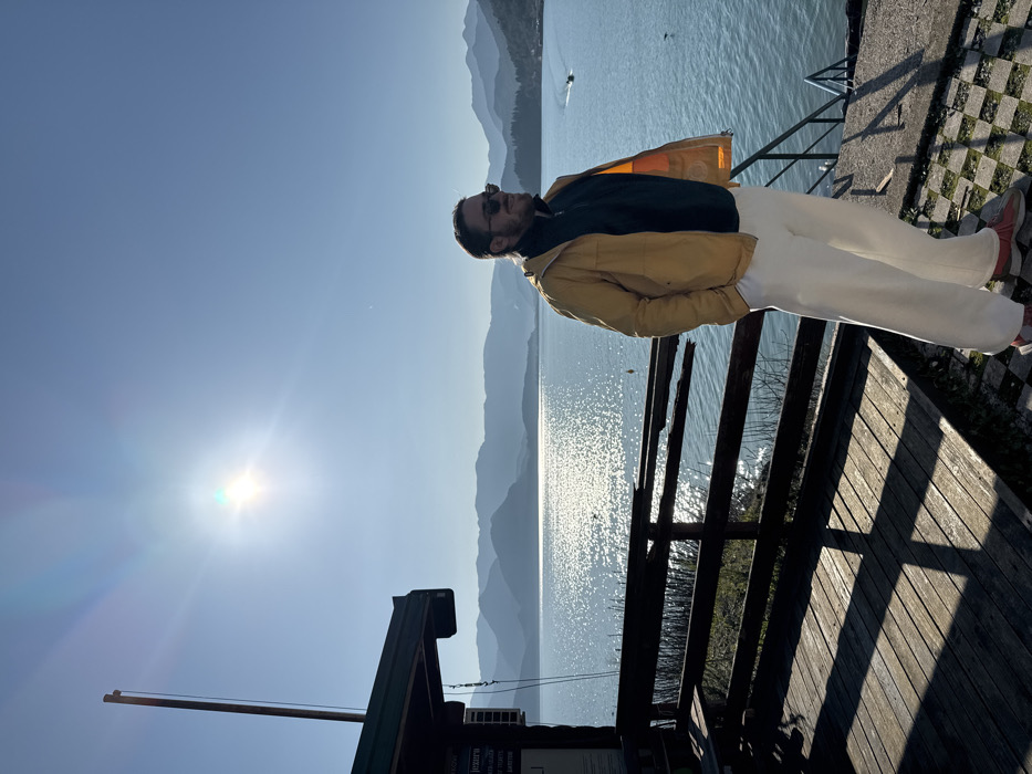 An outdoor scene captured on a sunny day in Sotonići, Montenegro, depicts a male subject standing near a body of water with mountains in the background.
The male subject, positioned on the right side of the frame, appears to be in his 20s or 30s, with light hair and a beard, wearing sunglasses. He is dressed in a mustard yellow hooded jacket, a dark blue or black half-zip long-sleeve shirt, light-colored (off-white) wide-leg pants, and sneakers with red detailing. His hands are in his jacket pockets, and he faces right, gazing horizontally.
To the left of the subject, a wooden deck or pier is visible, featuring a weathered wooden railing separating it from the water. Shadows from the railing and an unseen structure are cast prominently across the deck. A small, dark-roofed structure, possibly a boathouse or shelter, is situated on the far left, with a flagpole extending vertically from its side. A partially obscured sign is affixed to the building's wall. Adjacent to the male subject on the right, metal stairs descend into the water. The ground beneath the subject is a checkered pattern of light and dark squares.
The central feature of the background is a large body of water, shimmering brightly from direct sunlight reflected on its surface. Layers of mountains, appearing blue in the distance, form the horizon line. A small motorboat is visible on the water's surface in the mid-right distance. A yellow buoy is present in the water closer to the left. The sky is clear and bright blue, with the sun prominently visible in the upper left quadrant, creating a lens flare effect. Sparse vegetation is visible between the deck and the water's edge.