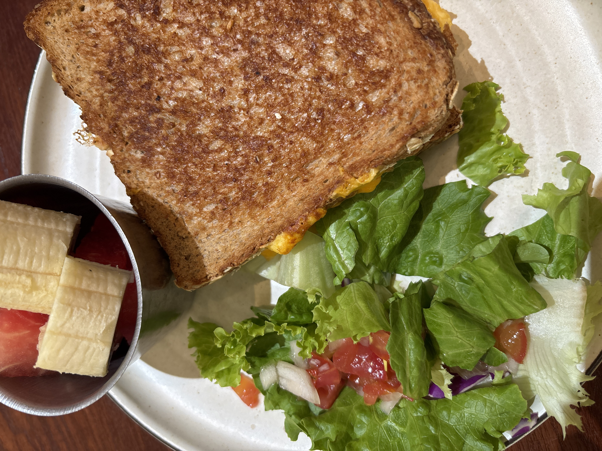 A grilled cheese sandwich is positioned on a plate next to a green salad with visible tomato and onion pieces, accompanied by a metal container of sliced banana and watermelon. The plate is on a wooden surface. There is no information about the location in the city of Đà Nẵng, Vietnam. No people, actions, or interactions are visible in this image.