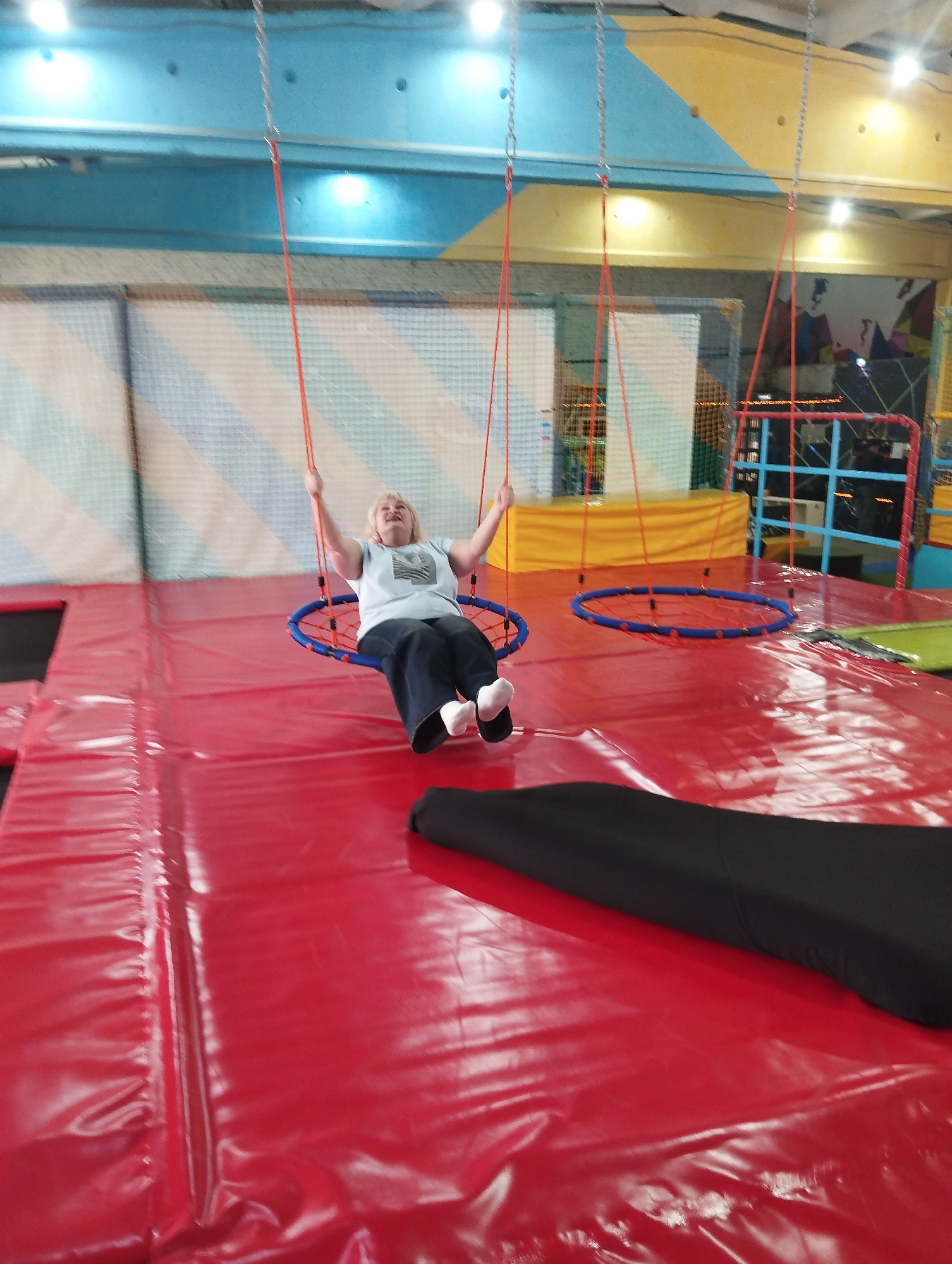 A woman sits on a blue-and-red netted swing in an indoor playground. The playground has red padded flooring and a background of striped safety netting, various play structures, and colored walls. The location is an indoor playground possibly in Votkinsk, Russia.