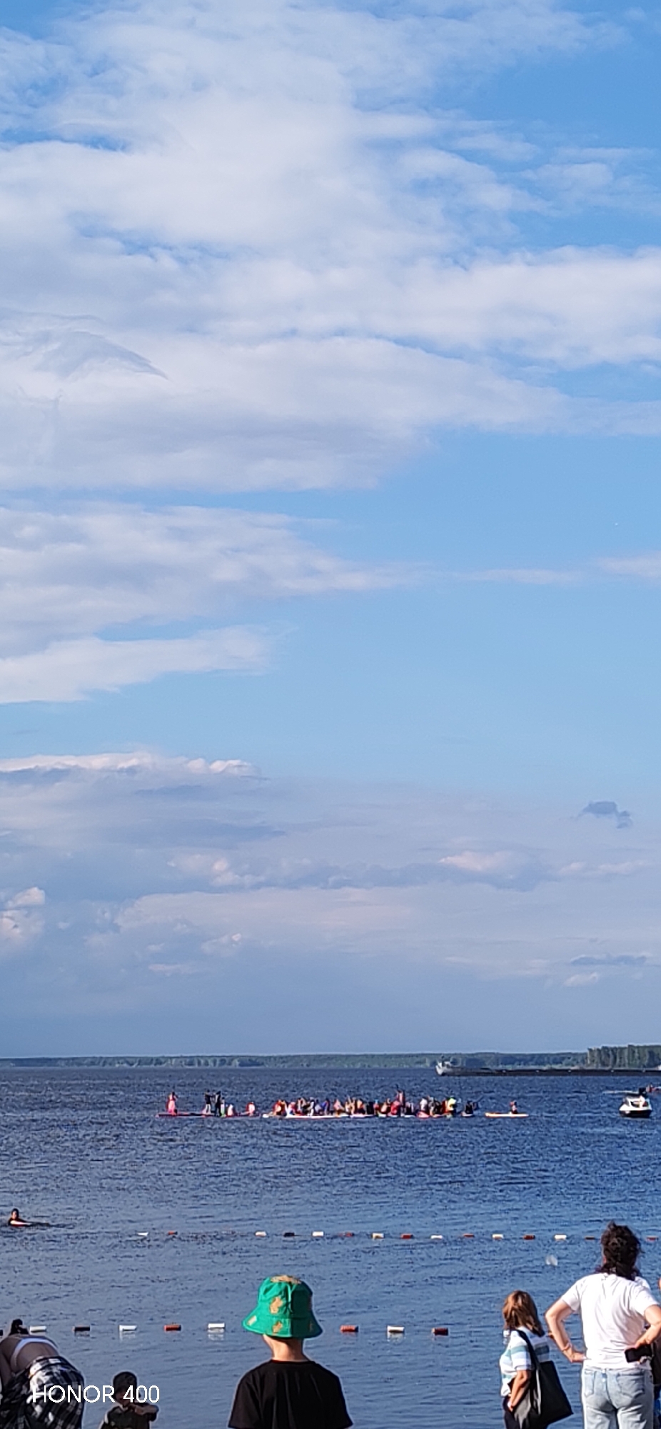 This media file captures a daytime scene on a body of water, likely a river or lake, in Chaikovsky, Russia. The sky is partly cloudy with a mix of blue sky and white clouds, suggesting fair weather. The water is calm, with a visible shoreline in the distance lined with trees.
The main subjects are a group of people participating in a water activity, possibly a race or organized event. They are spread across a long line of what appear to be paddleboards or kayaks, many of them standing. The group is quite large, suggesting a communal gathering or sporting event. Some individuals are also visible on the shore in the foreground, watching the activity on the water. These include a young person wearing a distinctive green bucket hat and other adults. One person is seen swimming or paddling in the water to the left of the main group.
In the background, a barge or large boat is visible on the water, adding to the sense of activity on the waterway. The overall mood appears to be one of a pleasant, outdoor event. The time of day is clearly daytime, given the bright lighting and the blue sky. The presence of a beach area with markers in the water indicates a recreational zone.
The text "HONOR 400" is visible in the bottom left corner of the image, indicating the device used to capture the photograph.