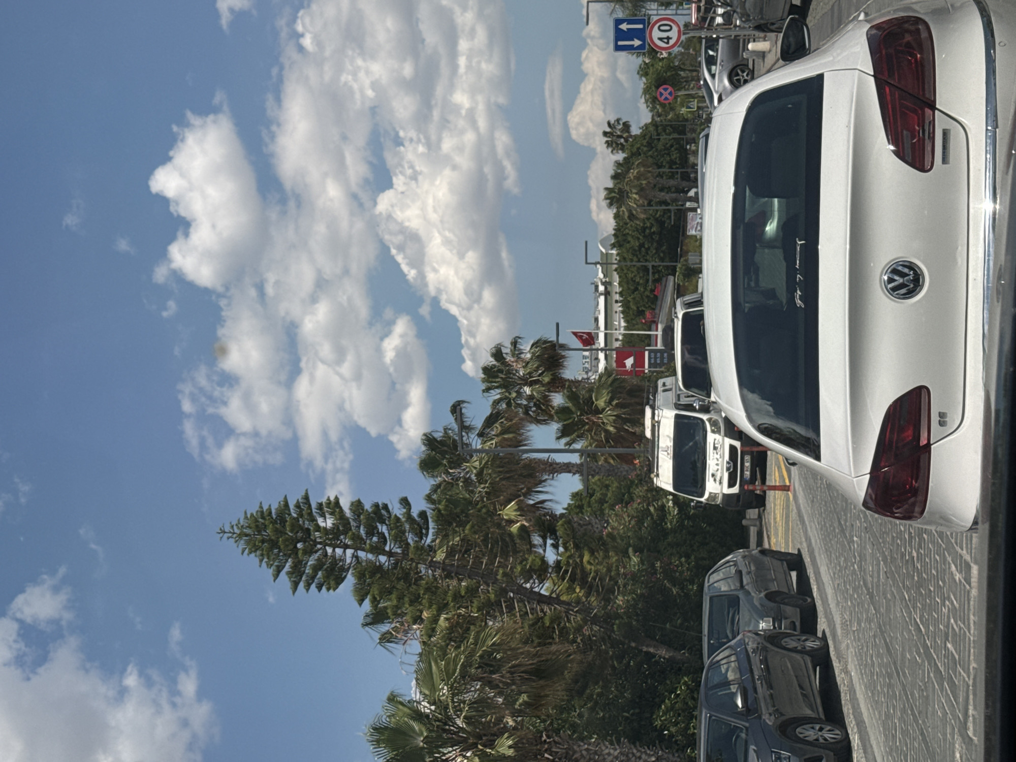 The image shows a street scene in Konyaaltı, Turkey, on a sunny day with a blue sky and scattered white clouds. Traffic is present, with a white Volkswagen car prominently positioned in the foreground, partially obscuring the view of other vehicles. A speed limit sign indicates 40 km/h. Palm trees and other greenery line the street, and the Turkish flag can be seen in the background. The perspective is from within a vehicle, looking out onto the street.