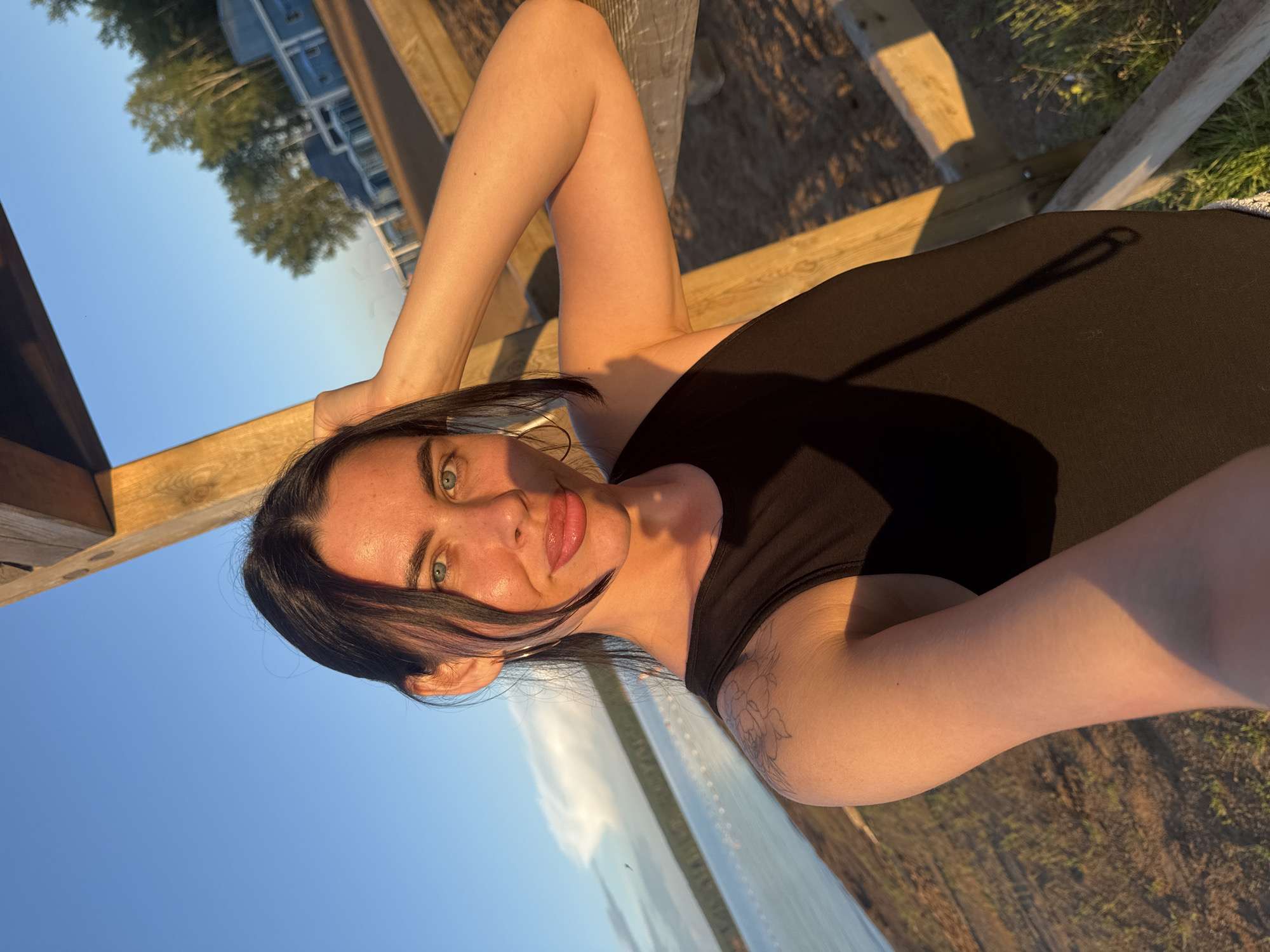 A selfie taken outdoors during golden hour shows a woman with dark hair and blue eyes. She is wearing a black sleeveless top and has her right hand on the back of her head. She is leaning against a wooden structure. The background shows a body of water, trees, and buildings, suggesting a lakeside or coastal environment. The lighting indicates it is either sunrise or sunset. The presence of buildings and a body of water is consistent with the landscape of Chaikovsky, Russia, a city located on the Kama River.