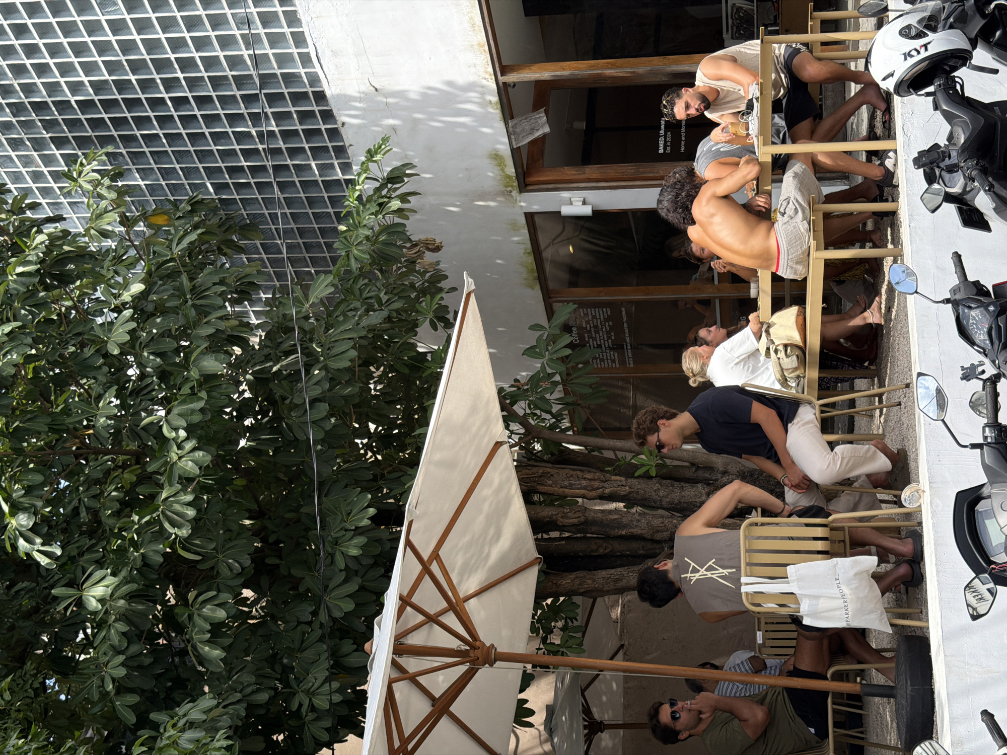 The image depicts an outdoor cafe scene in Kuta Selatan, Indonesia. A large, leafy tree occupies the left side of the frame, partially obscuring a building with a grid-like window design. An open umbrella with a white canopy and wooden frame is positioned in the foreground, providing shade to the seating area. Several people are seated at the cafe tables, some engaged in conversation, while others appear to be observing the surroundings. A row of parked motorbikes is visible on the right side of the frame. A sign for "BAKED. Uluwatu" and "BAIED BREW BAR AND ROASTERY PADONAN" can be seen indicating the cafe's name and offerings. Another sign includes “@PARKERPEOPLE_”.
