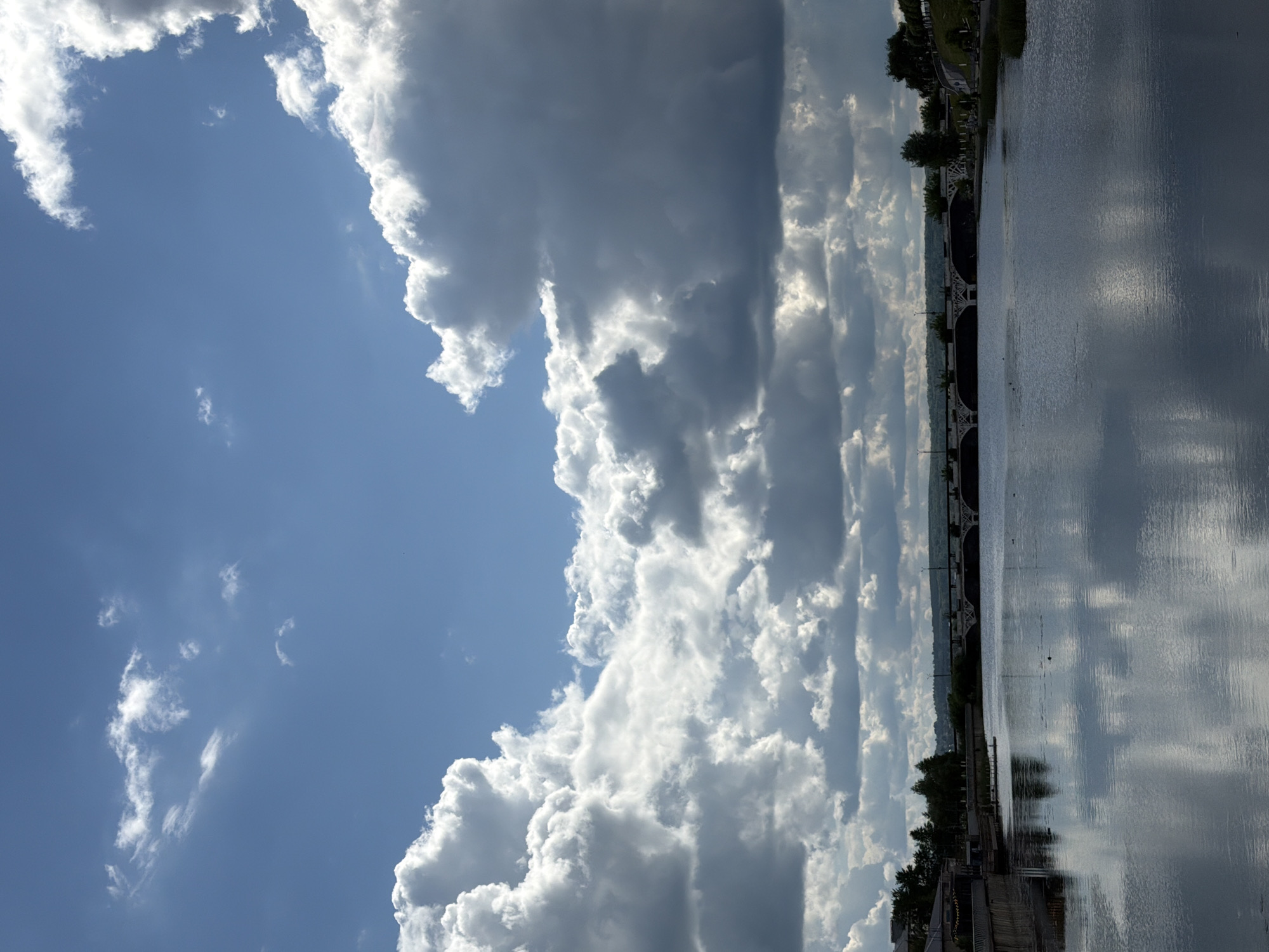The image shows a vertical view of a body of water with a bridge in the distance, under a sky dominated by clouds. The clouds range from bright white to dark grey and fill the majority of the frame. The water reflects the sky, with variations in the reflection due to surface conditions. The bridge appears as a dark structure spanning the water. Vegetation is visible on the bank of the water. Based on the location information (Votkinsk, Russia), it is possible that the body of water is the Votkinsk Reservoir. However, without further details, precise identification of the location and objects is not possible.