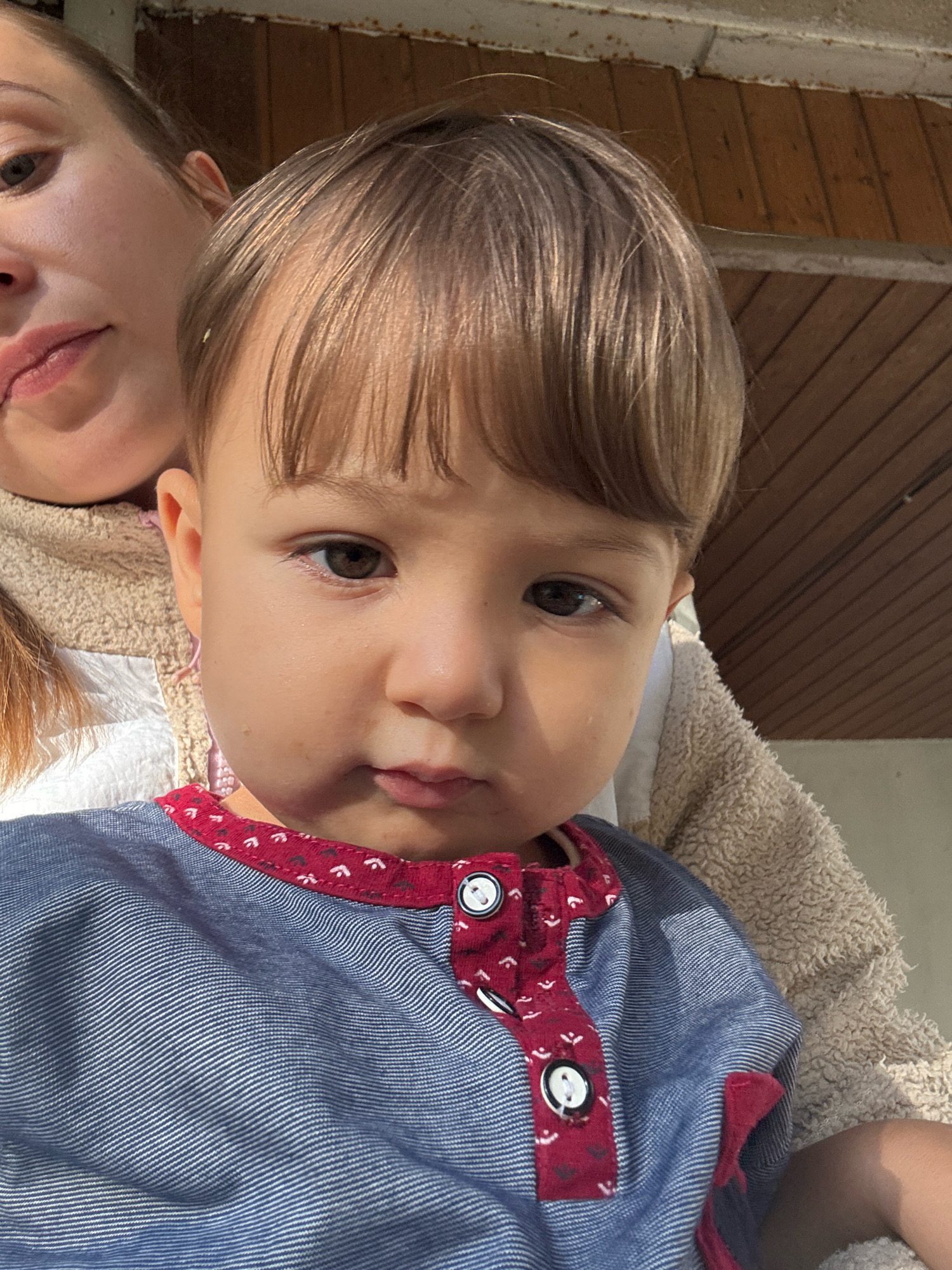 A medium shot captures a close-up of a young child, likely a toddler, being held by an unseen adult. The child's face is the primary focus, with large, brown eyes looking directly at the camera. The child has fair skin and light brown hair cut in a bob style. The child is wearing a blue shirt with thin white pinstripes and a red fabric placket adorned with a pattern of small white shapes. Two white buttons are visible on the placket. The adult's shoulder and part of their face, including an eye and lips, are visible on the left side of the frame. The background appears to be an outdoor setting with wooden paneling, possibly the side of a building or a fence. The lighting suggests it is daytime, with sunlight casting shadows. No specific location in Überherrn, Germany is identifiable from the visual information.
