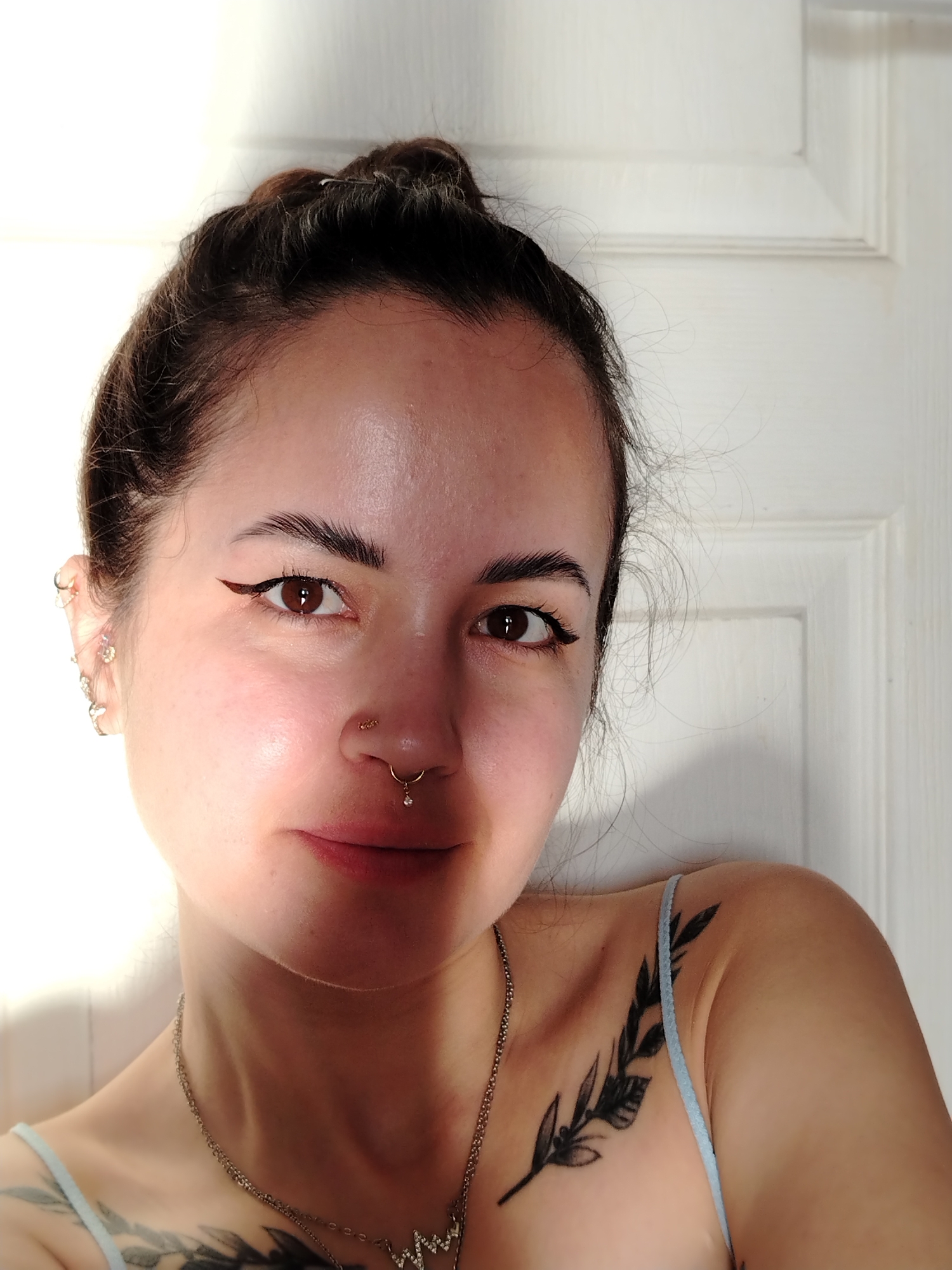 A close-up portrait shows a fair-skinned woman with brown hair pulled into a messy bun. She has dark eyebrows, brown eyes with eyeliner, and a nose piercing. She wears a light-colored tank top, a silver chain necklace, and has tattoos on her chest and shoulder. The background is a white door. The city of Noviy, Russia is not featured.