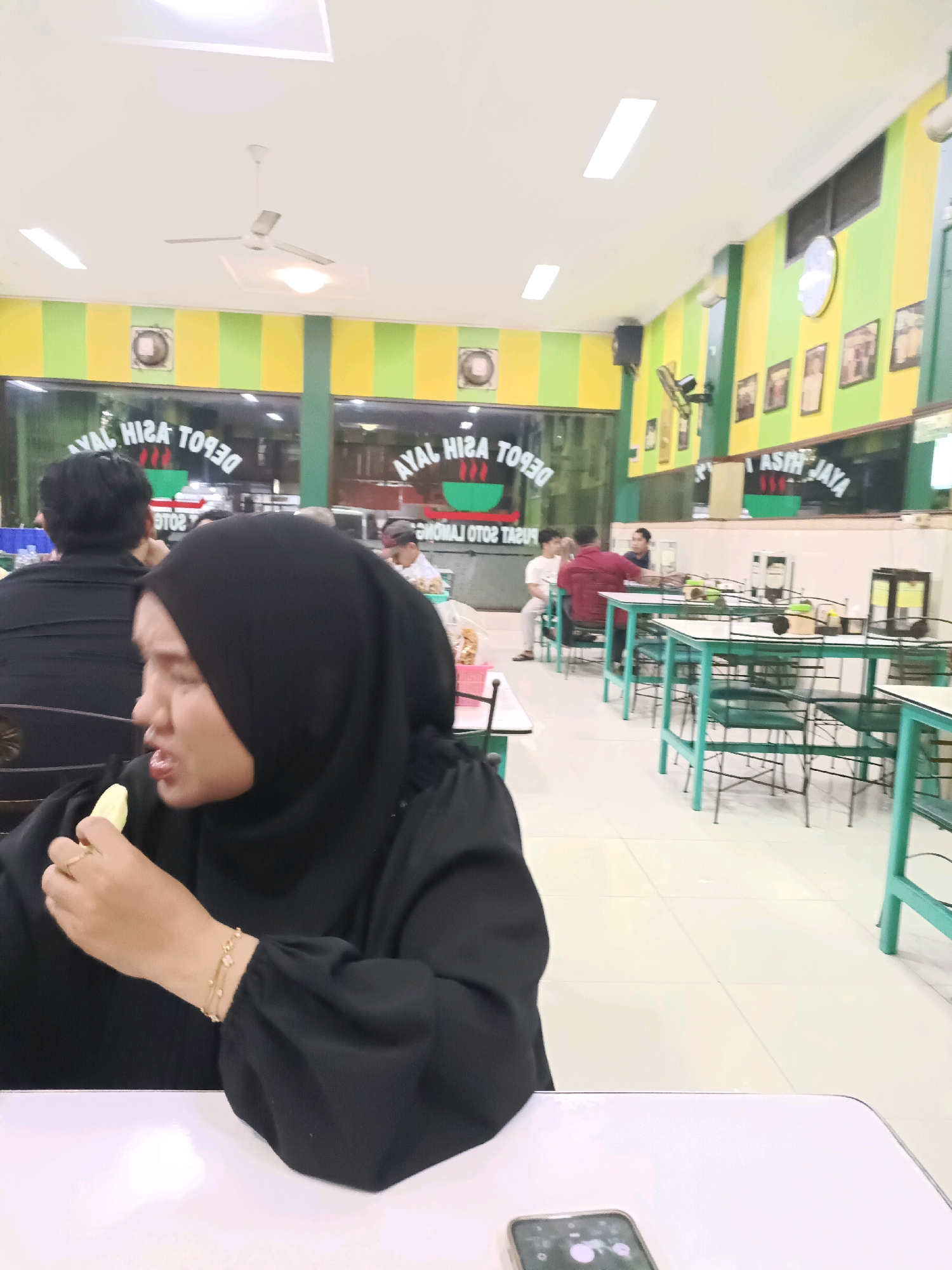 A medium shot captures a woman wearing a black hijab and dress, seated at a white table in what appears to be a restaurant in Dapur, Indonesia. She is shown in profile, looking to her left and eating a piece of food held between her fingers. A thin gold bracelet with charms adorns her left wrist. In the background, other patrons are visible, seated at tables. The restaurant features walls painted with alternating green and yellow stripes, and a ceiling fan is visible overhead. Several framed pictures hang on the wall. Tables with green metal frames and white tops, accompanied by chairs, are arranged throughout the dining area. The flooring is tiled.