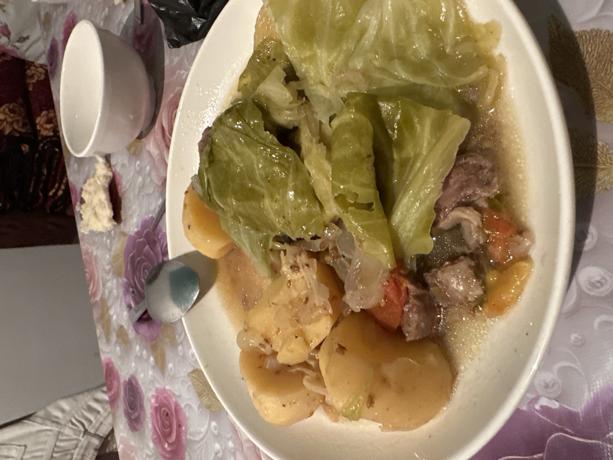 In an indoor scene with a floral tablecloth, a white plate holds a meal consisting of stewed cabbage, potatoes, and meat chunks with tomatoes. A spoon rests on the edge of the plate. A white bowl and a piece of bread are visible on the table to the right of the plate. The background shows upholstered furniture and a wall.