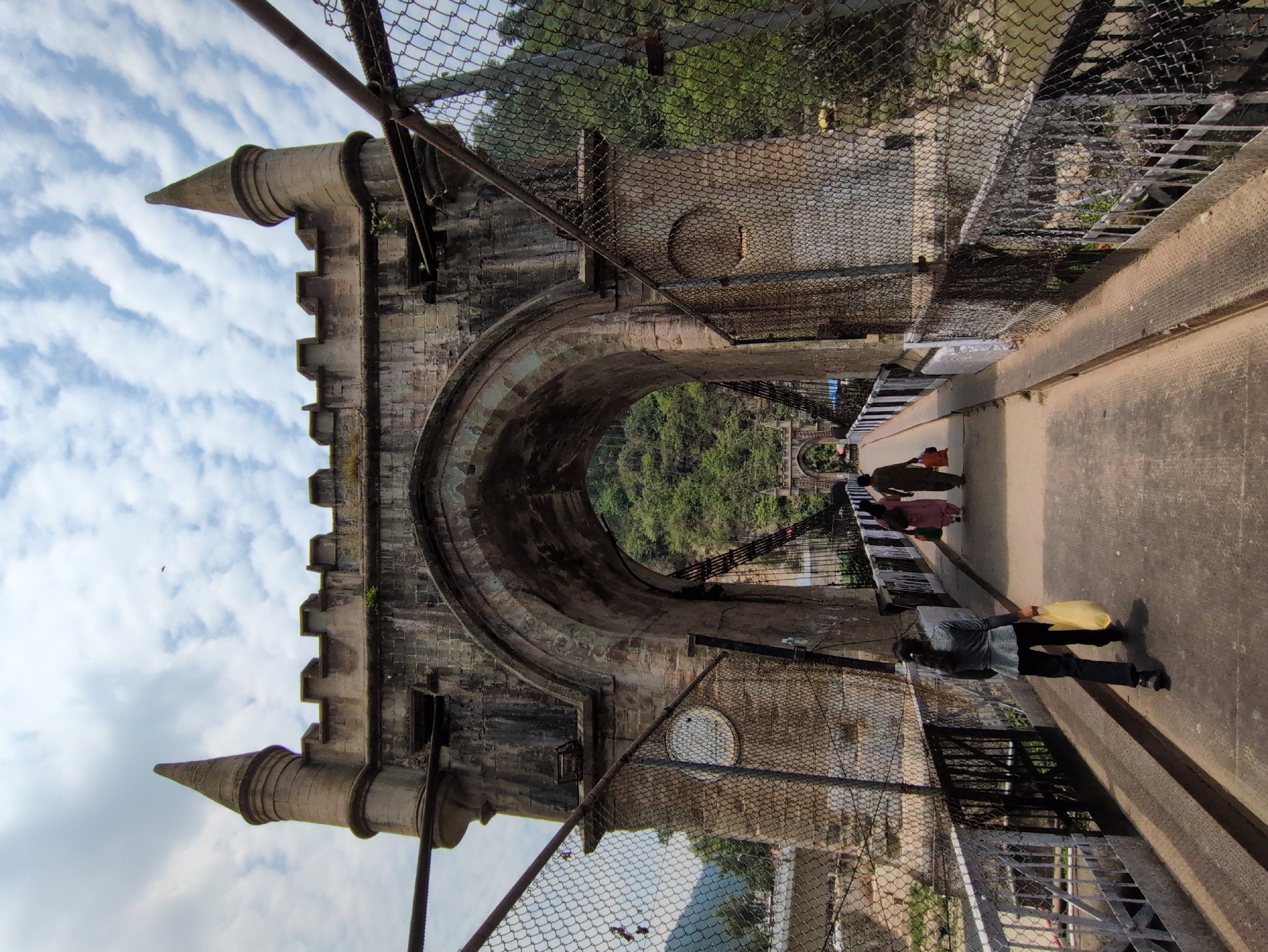 The image shows a stone bridge with a large archway in the city of Mandi, India. The bridge has a suspension structure with wire mesh fences on either side. Two women and one person, identified by the visible leg and shoes, are walking across the bridge. The woman in the foreground is carrying a yellow bag. The bridge appears to be old, with some signs of wear and tear on the stone structure. The background shows lush green hills and a cloudy sky. The bridge is likely the Victoria Bridge, a historic landmark in Mandi, known for its distinctive architecture.