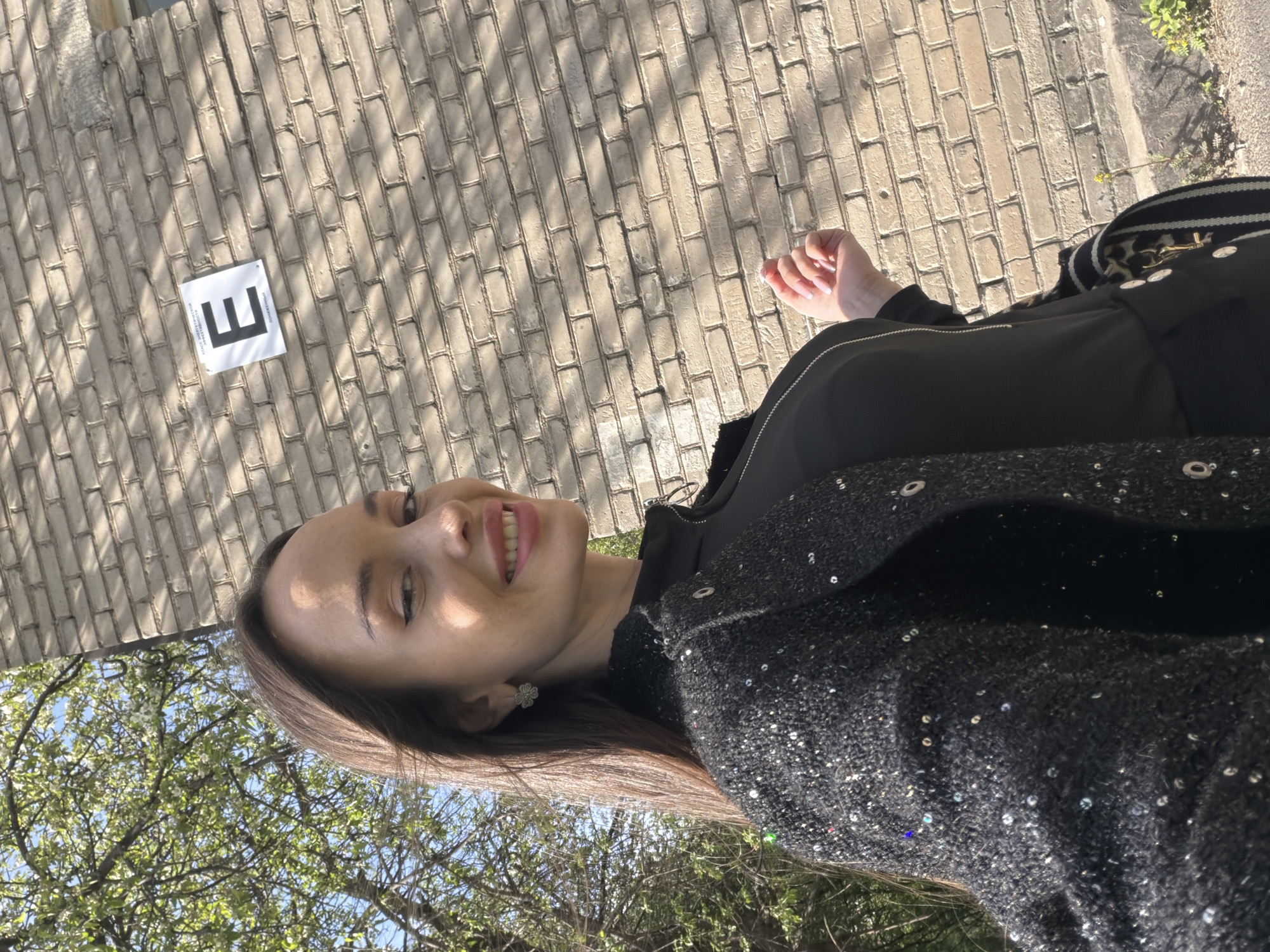 The image is a rotated selfie of a woman standing outside on a sunny day, possibly in Saint Petersburg, Russia. She is smiling, wearing a black jacket with a sequined or sparkly outer layer, and has long brown hair. A brick walkway or road is visible in the background, with a sign displaying the letter "E" attached to it. Trees with green leaves and blue sky are visible in the lower left corner. A hand is visible on the right side, and a black and white striped strap with gold hardware suggests a bag is also present.