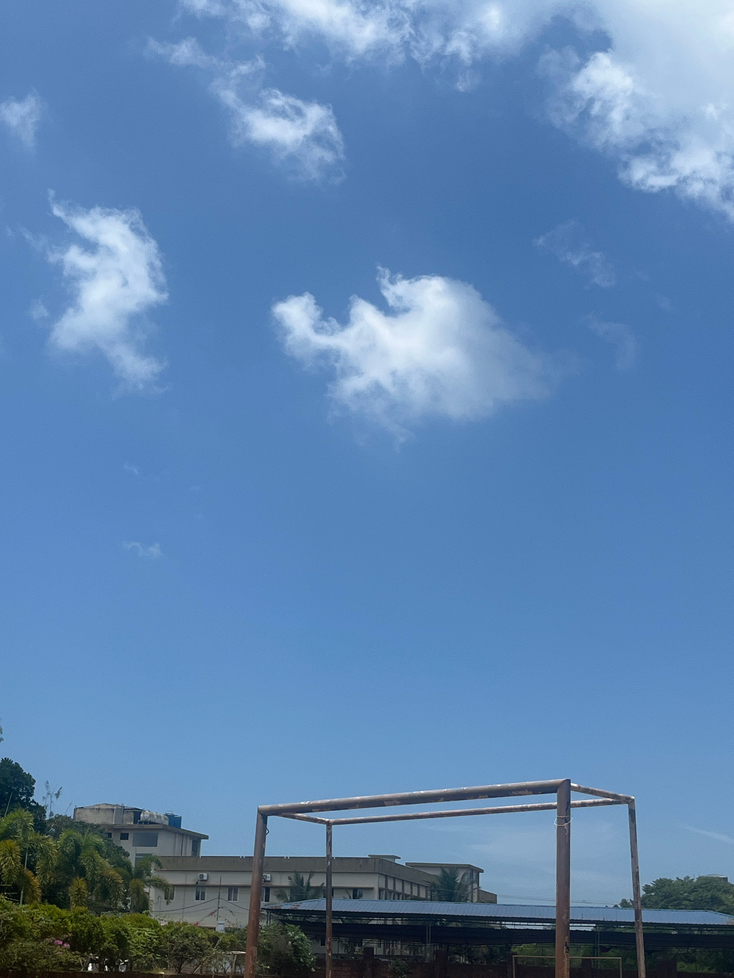 The image shows a bright, clear blue sky with scattered white clouds. In the lower part of the frame, there is a partially visible structure, likely part of a sports field or playground, with rusted metal poles and crossbars. Beyond this structure, buildings and lush green trees are visible, suggesting an outdoor urban or semi-urban setting in Malappuram, India. The buildings appear to be multi-story structures, some with balconies and air conditioning units, consistent with architecture found in the region. The overall scene conveys a sunny day. There are no people or discernible actions taking place.