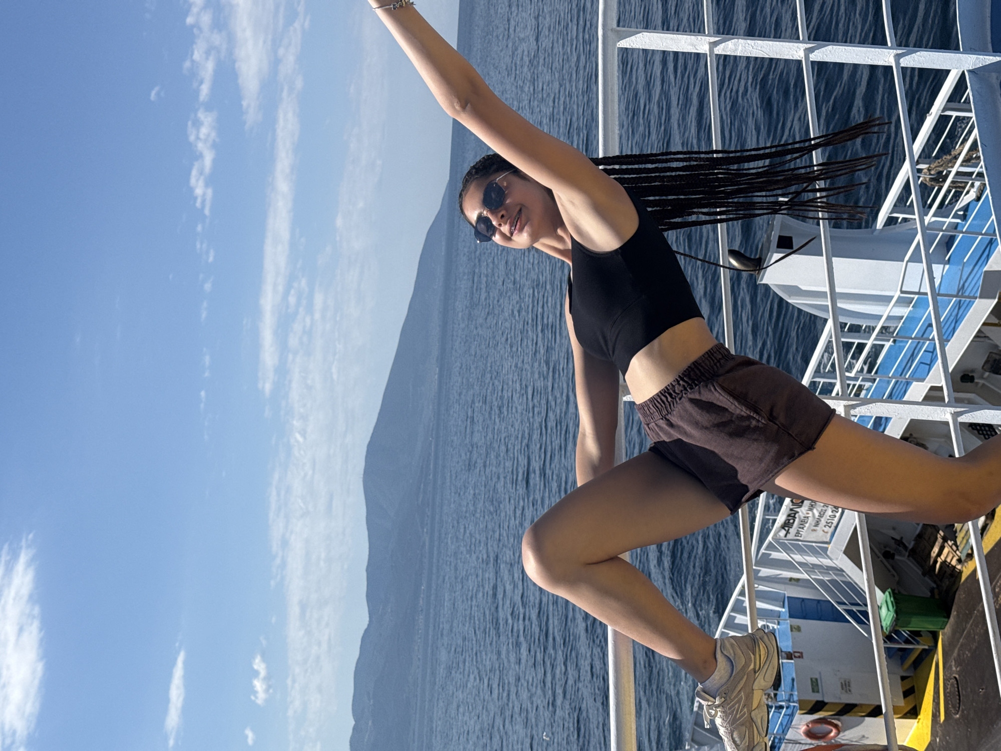 A young woman with braided dark hair and sunglasses leans back against a railing on a ferry, her left leg extended upwards. She wears a black crop top and brown shorts. The ferry is sailing on a blue sea under a clear sky with scattered clouds. In the background, a mountainous coastline is visible. The ferry deck features a green trash can and other nautical equipment. The image appears to have been taken in Greece.