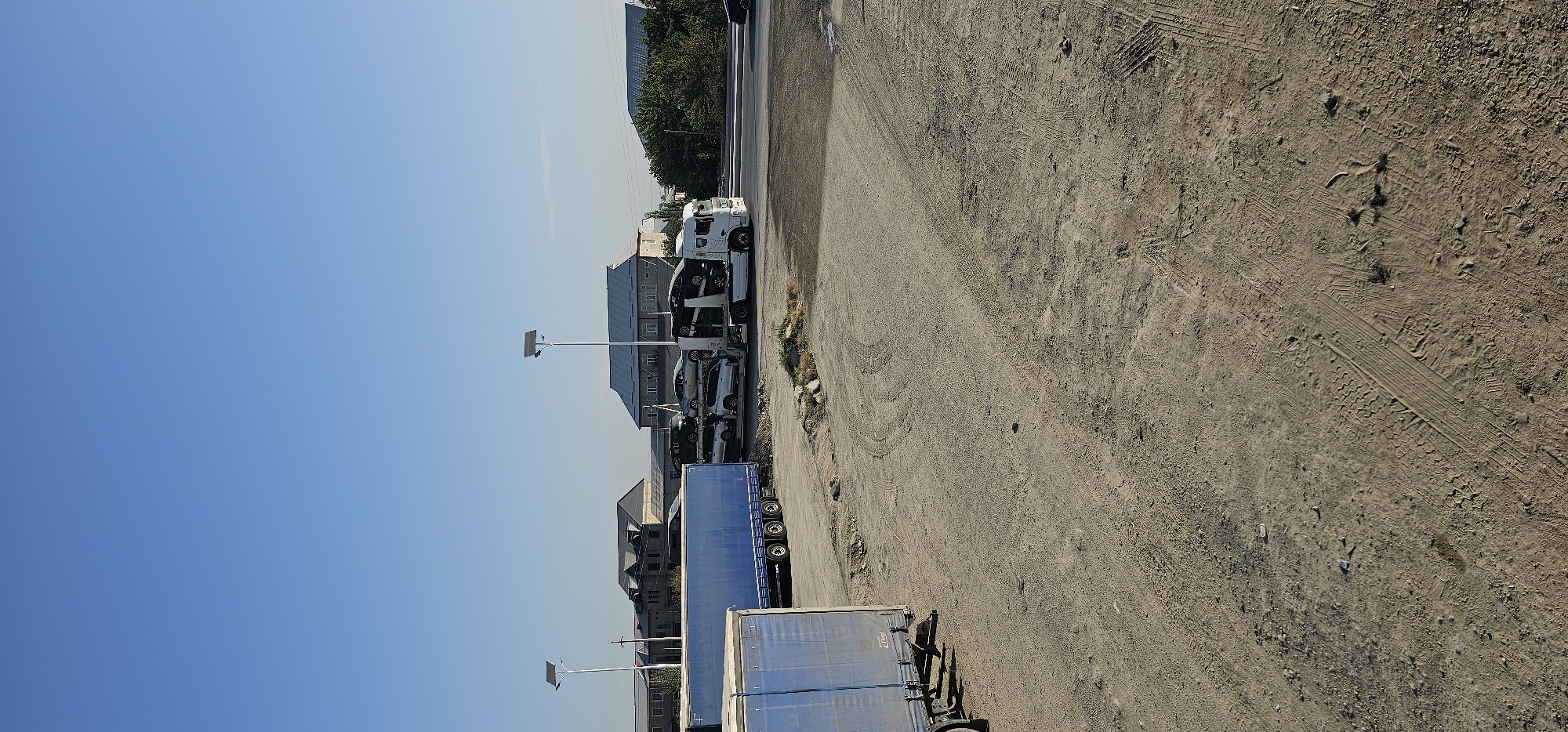 The image depicts an outdoor scene in the city of Sheyhancha, Uzbekistan, under a clear blue sky. In the foreground, a dusty, gravelly ground area is visible, marked with tire tracks from vehicles. To the left, a blue truck trailer with multiple axles is parked. In the middle ground, a white car carrier truck is parked, loaded with several cars. Behind the truck, there are buildings with peaked roofs, possibly residential or commercial structures, and two tall lamp posts with solar panels are present. A road is visible in the background, with some trees and vegetation to the right. The scene appears to be a parking or staging area for vehicles.