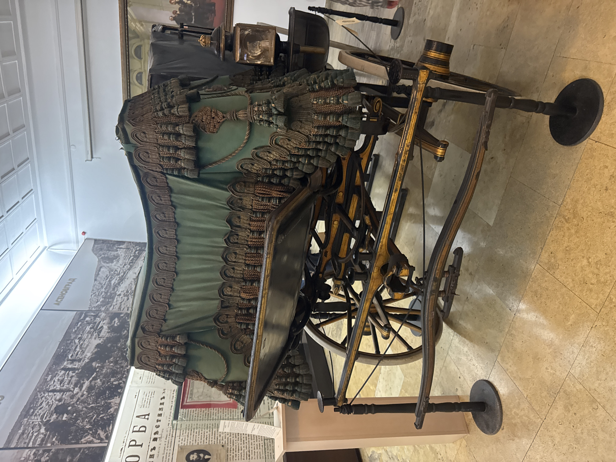 The image depicts an antique horse-drawn carriage on display in a museum. The carriage has a green cloth roof with decorative trim. The frame and wheels are made of dark metal with gold detailing. In the background, there's a display with historical documents and images, one with the word "АСЕОНГАД" and another labeled "ОРБА" visible. The carriage is positioned on a light-colored tile floor, inside what seems to be an exhibition hall. The photograph was taken in Plovdiv, Bulgaria.