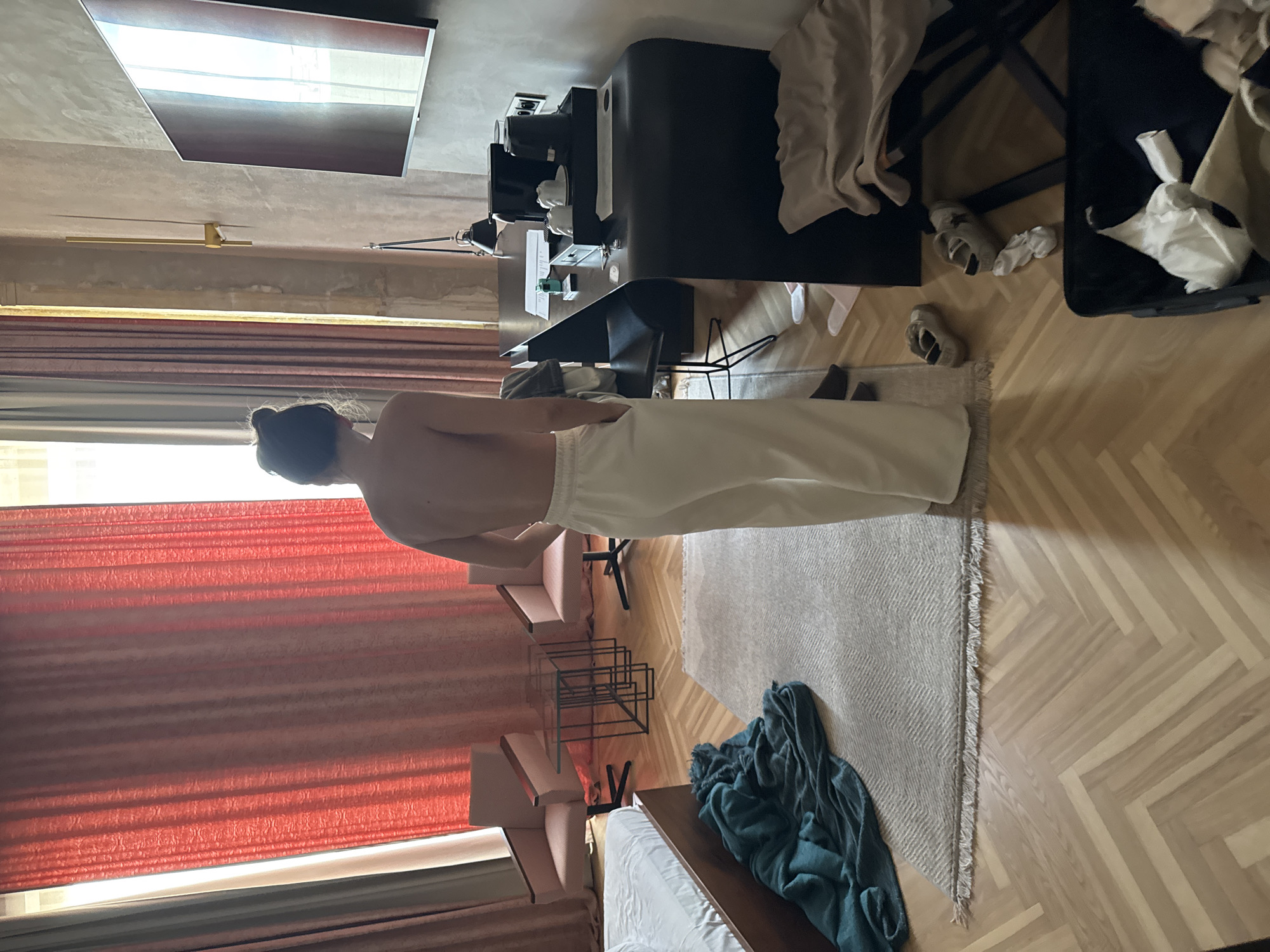 The image depicts an indoor scene, likely a hotel room, in the city of Unknown, Unknown. A single person, seen from the back, stands slightly left of center in the foreground. The individual is topless and wears light-colored, possibly white or off-white, wide-leg trousers with hands in the pockets. Their dark hair is gathered in a bun.
The room features light-colored wooden parquet flooring laid in a herringbone pattern, partially covered by a light grey rug with a textured weave and fringed edges. To the left, a section of a bed with a white mattress and a dark wooden frame is visible. A dark teal or green blanket lies crumpled on the floor beside the bed.
In the mid-ground, a pair of large windows are draped with floor-length, textured, reddish-orange curtains. A sliver of an outer, lighter-colored curtain and bright daylight are visible on the far left. In front of the windows, two light pink or beige armchairs with dark wooden armrests and black swivel bases face each other. Nested between them are two side tables with glass tops and dark, geometric metal bases.
On the right side of the room, a dark, modern desk with a curved front is positioned against a textured, light brown or grey wall. A black desk chair is tucked beneath it. The desk surface holds a black coffee machine, a black electric kettle, white cups on a tray, various papers, and a black desk lamp. Above the desk, a flat-screen television is mounted on the wall, reflecting light from the window. A gold-toned, linear wall sconce is visible higher on the wall.
Near the floor on the far right, an open dark suitcase or bin contains crumpled white clothing. On the floor nearby are two pairs of light-colored sneakers, a crumpled white sock, and two pairs of white slippers. A beige throw or garment is draped over an unseen item near the suitcase. The natural light from the windows creates bright reflections and shadows on the floor and the television screen.