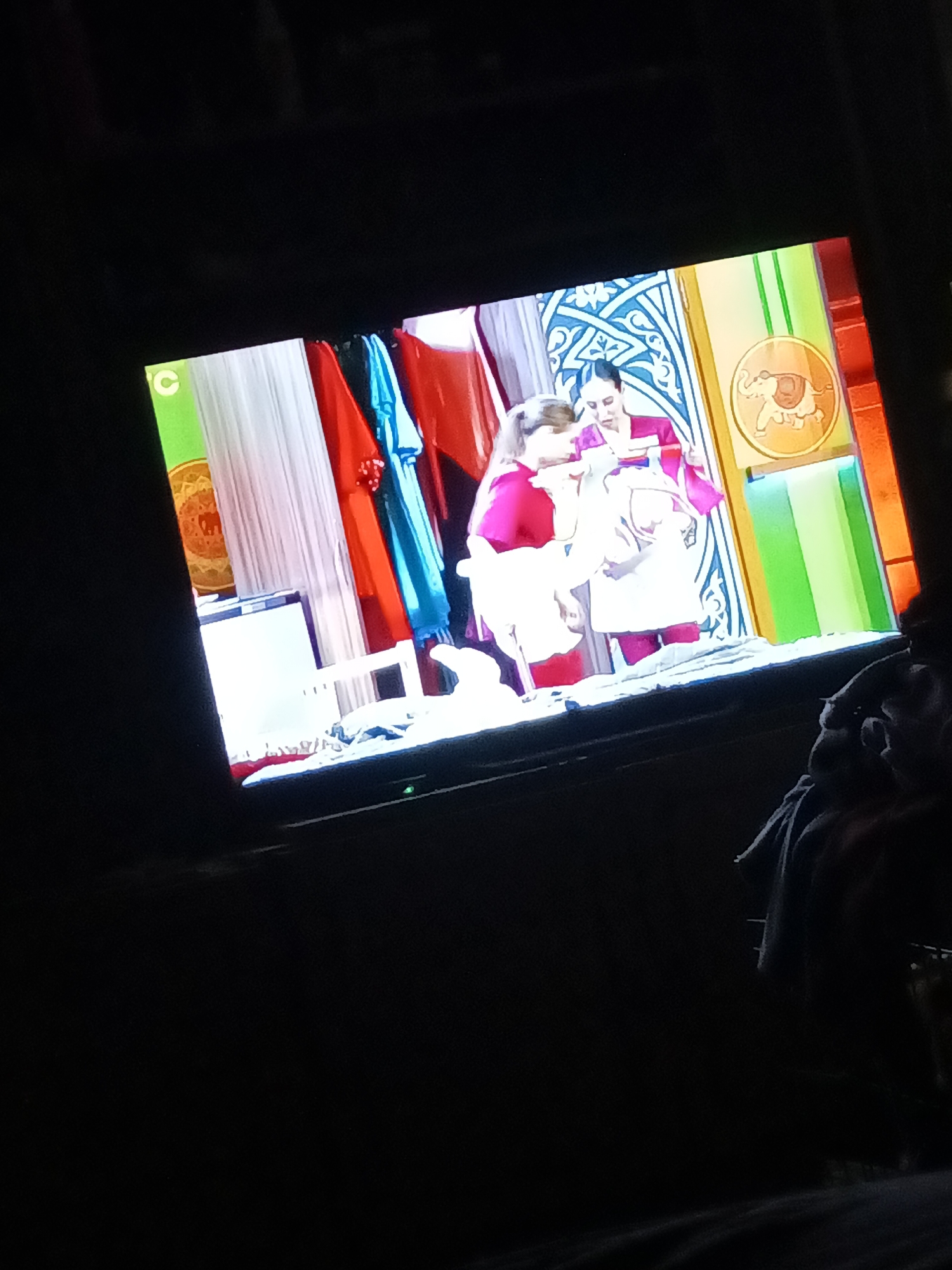 The image shows a television screen displaying a scene with two people, likely children, dressed in colorful clothing. The person on the left wears a bright pink top and white apron, while the person on the right wears a pink top and a white apron with pink trim. They appear to be engaged in an activity. The background on the screen is a set with vibrant colored panels, including green, red, blue, and white. There is also a decorative element with a blue pattern and an image of an elephant in a circular frame. The television is positioned in a dark room, with a person's shoulder and head visible in the foreground on the right, suggesting the image was taken from a spectator's perspective. The lighting is dim, with the primary light source coming from the television screen. The location in the city of Новопетровское, Russia, is not discernible from the image.