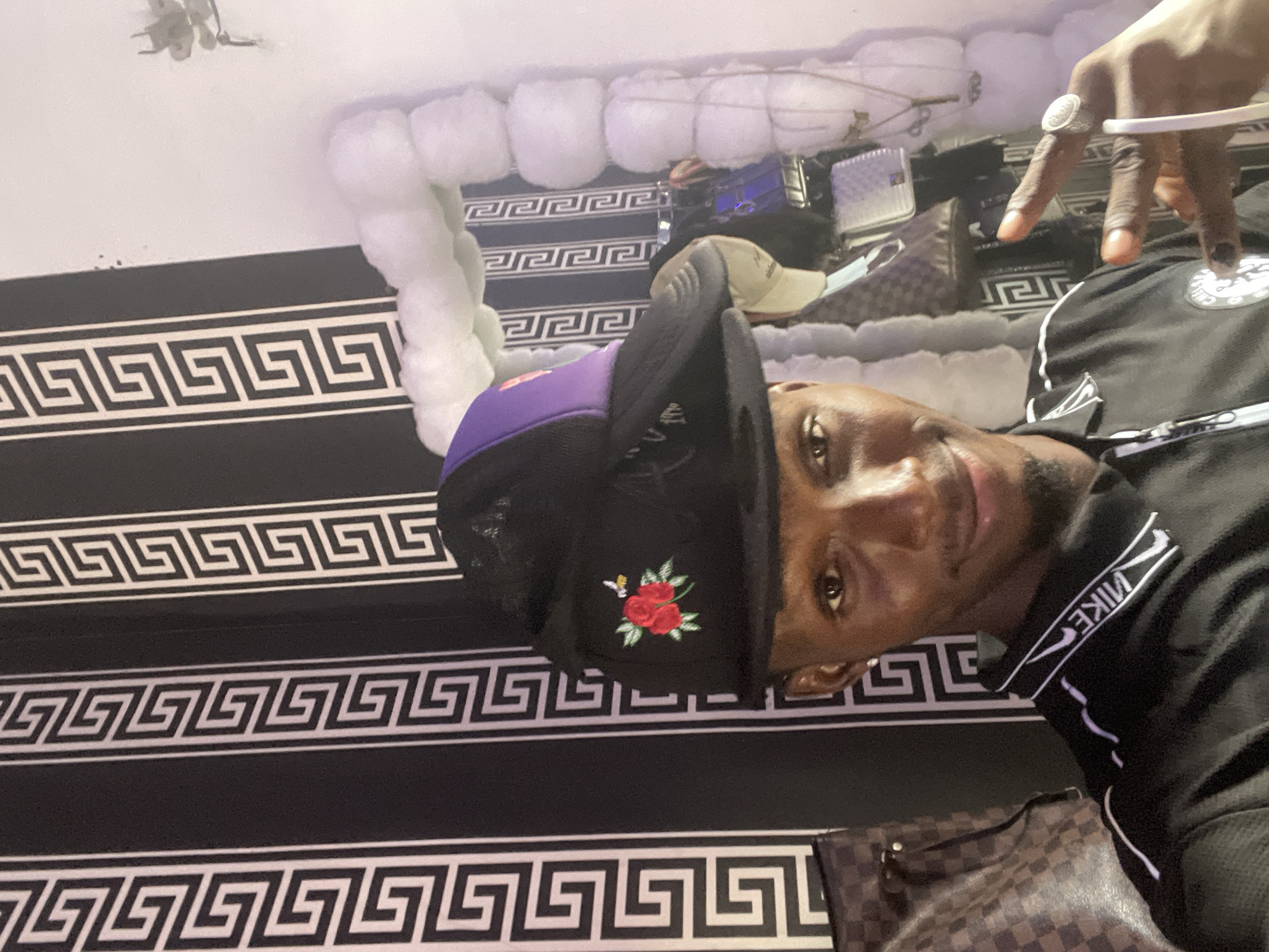 A man is indoors in a room with black and white patterned wallpaper. He is wearing a black and purple baseball cap with a red rose design, and a black shirt with white Nike branding. He is flashing a hand sign with a silver ring. There is a white, cloud-like decoration on the ceiling, and a collection of handbags and other items are arranged behind him. It is not possible to confirm the location is Lokoja, Nigeria from the image alone.