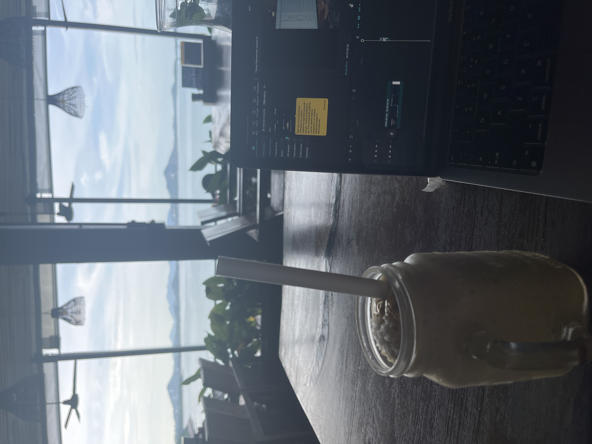 This media file shows a close-up of a glass jar filled with a light-colored beverage, possibly a smoothie or milkshake, with a straw inserted. The jar is placed on a dark wooden table. In the background, through a large window, a scenic view of a body of water with islands and hills under a cloudy sky is visible. There are also glimpses of indoor elements, such as hanging lamps with a woven design and lush green plants. A laptop is open on the table, displaying what appears to be video editing software with Russian text. A yellow sticky note is attached to the laptop screen.
The text on the laptop screen and sticky note indicates video editing activity. The Russian text translates to instructions about a "split scene" function available in the software, suggesting the user is actively working on a video project. The timecodes visible on the screen (e.g., 00:06:50:14) further confirm video editing. The overall setting appears to be a cafe or workspace with a view of a tropical landscape, likely during the day given the natural light. The mood seems to be one of focused work in a relaxed, picturesque environment.
