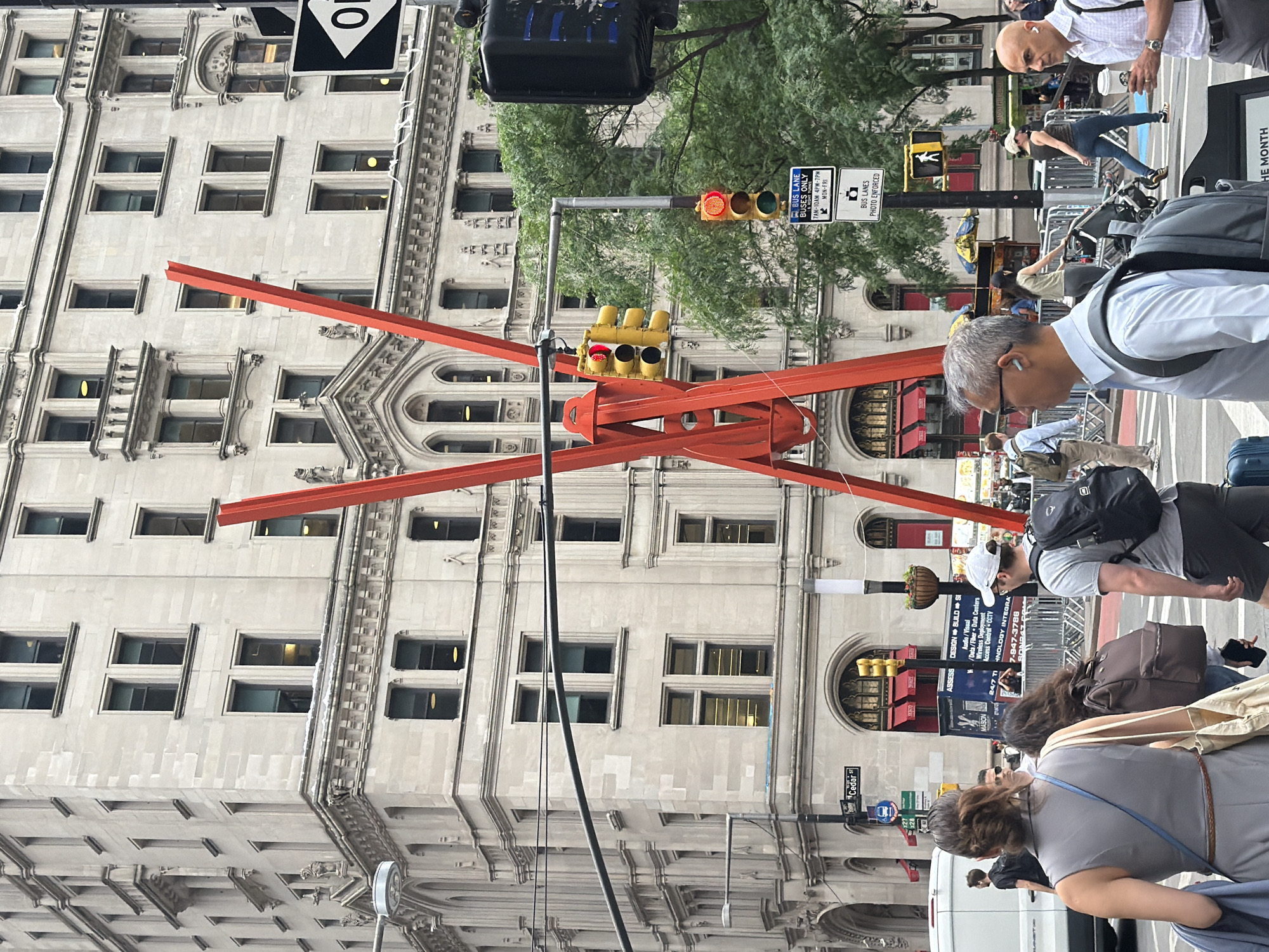 This is a street-level, eye-level shot taken in a busy urban environment, likely New York City, given the visible street signs and architecture. The scene is dominated by a large, ornate stone building with numerous windows and detailed architectural features. In the foreground, a striking abstract sculpture made of bright orange beams is prominently displayed, intersecting the frame and creating a dynamic visual element.
Several people are present, walking and interacting, suggesting a normal day with pedestrian traffic. They are dressed in casual summer attire, and some are carrying bags, indicating they might be commuting or shopping. One man in the foreground is wearing a light blue shirt and glasses, looking to his right. Another man with a bald head and a white shirt is visible further back, holding a coffee cup.
Traffic signals are visible, with one showing a red light, implying that vehicles are stopped. Street signs like "Cedar st," "BUS LANE BUSES ONLY," and "BUS LANES PHOTO ENFORCED" provide specific location context and traffic regulations. The architecture of the building, with its classic design and arched windows, suggests a historical or financial district. The presence of trees and the overall brightness of the scene indicate it's daytime, likely under overcast skies given the diffused light. The general atmosphere appears to be one of everyday urban activity.