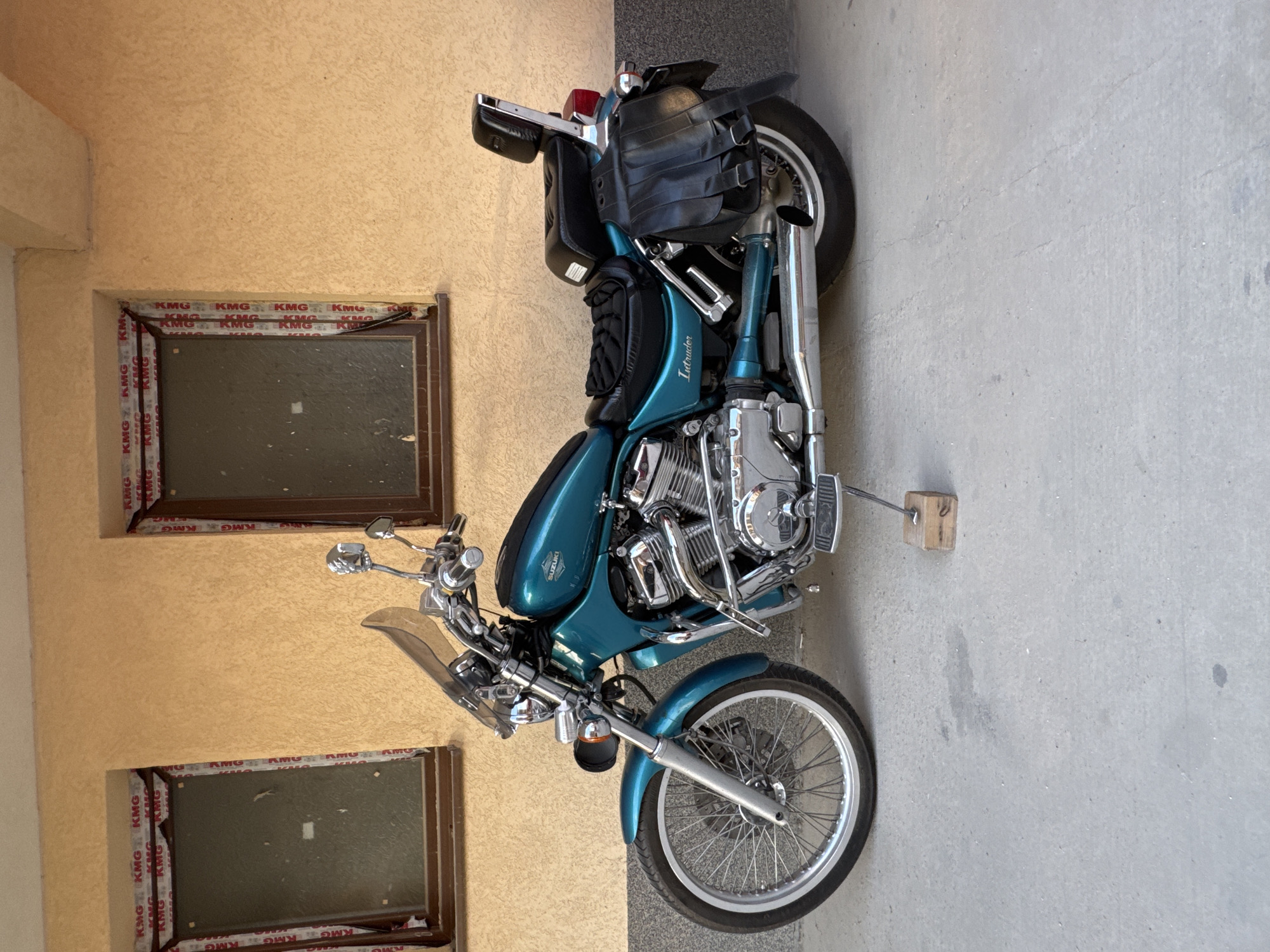 Here is a forensic description of the image:
Scene: The image shows a teal-colored Suzuki Intruder motorcycle parked outdoors next to a light orange stucco wall with two windows trimmed with brown and a KMG logo pattern, and on a concrete ground surface.
Objects:
- Motorcycle: A teal-colored Suzuki Intruder is the main subject. It has a windshield, chrome engine components, and black leather saddlebags attached to the rear.
- Wall: The stucco wall is light orange in color and has two rectangular windows. The windows are trimmed with brown edging that feature a repeated "KMG" logo pattern.
- Windows: The two rectangular windows are closed.
- Ground: The ground is made of concrete.
Environment:
- Outdoor setting, likely next to a building.
Inference about Location:
The presence of the Cyrillic script in the "KMG" logo around the window frame suggests that the location is likely in Bulgaria. As the prompt asked specifically for Plovdiv, we may infer that the scene is in that city.
---