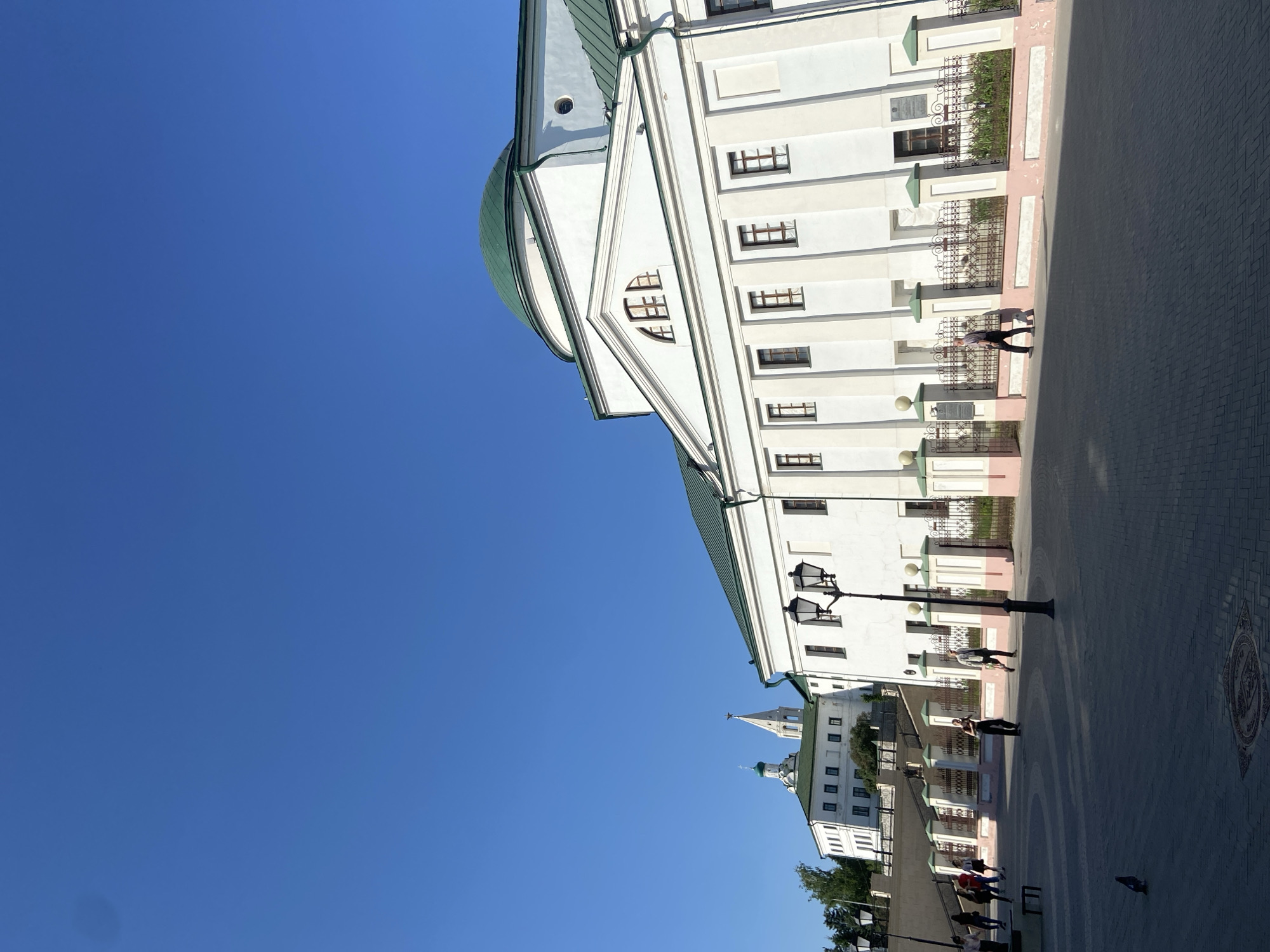 The image shows a view of Kazan, Russia, featuring white buildings with green domes set against a clear blue sky. People are walking along a paved area adjacent to the buildings. Street lamps are visible along the walkway. The architecture suggests a historical or cultural site within the city.