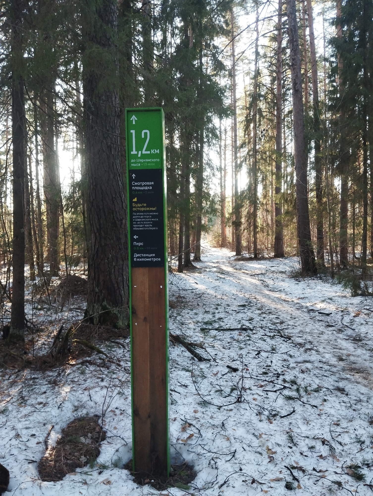 The image shows a snow-covered forest trail in Votkinsk, Russia. A green and brown signpost stands prominently along the path. The sign indicates that the path leads to various destinations, including a pier ("Пирс"), a viewing platform ("Смотровая площадка"), and Sharkansky Cape ("до Шарканского мыса"). The sign also includes a warning to "Be careful!" ("Будьте осторожны!"). The distance to Sharkansky Cape is marked as 1.2 km. The forest consists of tall, bare trees with the ground partially covered in snow and fallen branches. Sunlight filters through the trees, creating a bright and airy atmosphere.