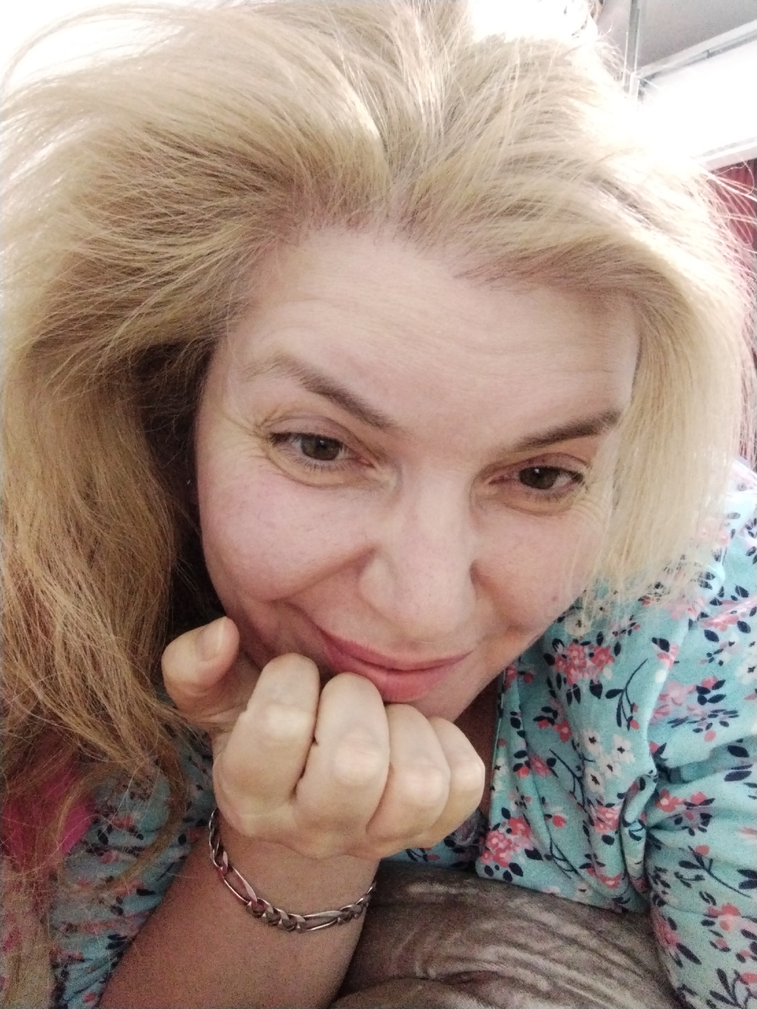 A close-up selfie captures a Caucasian woman with blonde hair and fair skin resting her chin on her hand. She wears a turquoise blouse with a floral pattern. The background is out of focus, with hints of a light-colored wall and possibly a ceiling fixture. The woman has a slight smile, and her right eye is visible, showing a brown iris. The lighting suggests an indoor setting. There are no identifiable landmarks or elements that would place the image in Al Mas'oudi, United Arab Emirates.
