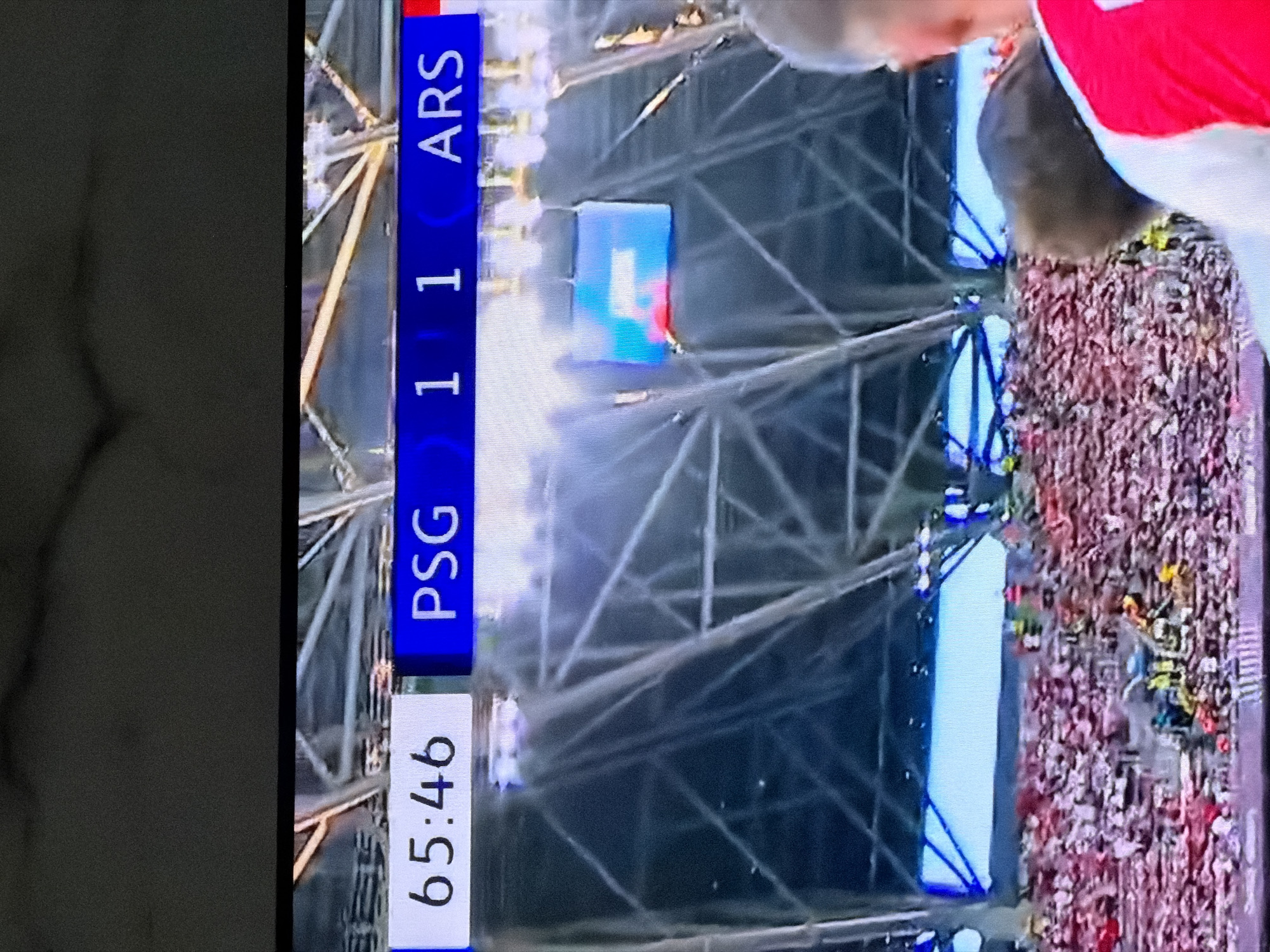 The image shows a soccer match score overlay of PSG 1 - 1 ARS, displayed at 65:46 of the game time. A portion of the crowd is visible, and floodlights are active. The location, Asenovgrad, Bulgaria, is not identifiable from the visual details of the image.