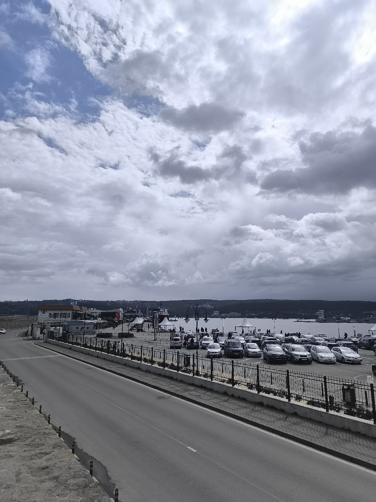 The image depicts a cityscape in Varna, Bulgaria, under an overcast sky with scattered clouds. A wide, gray asphalt road runs diagonally across the lower half of the frame, bordered on the right by a wrought iron fence and a stone wall. Beyond the fence is a parking area filled with numerous cars. In the distance, a body of water, possibly the Black Sea, is visible. On the waterfront, there are several small buildings, canopies, and people are seen walking along the pier. The horizon is marked by a dark, wooded area.