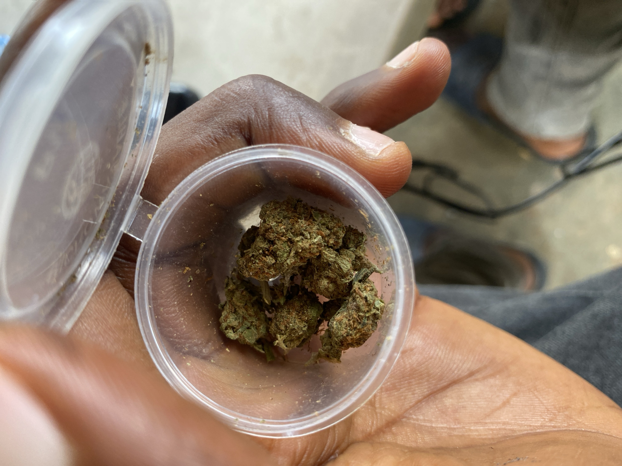 A close-up image shows a person with dark skin holding a small, clear, round plastic container with a hinged lid. The container is filled with several dried, green and brown plant buds, consistent with cannabis. The person's fingers and palm are visible, cradling the container. The lid of the container is open and partially visible at the top left. In the softly blurred background, another person's leg or foot, wearing dark footwear such as a slipper or sandal, is partially visible on a light-colored, possibly concrete, surface. Dark fabric, likely clothing, is also visible around the background foot. The lighting suggests daylight. The image appears to depict the presentation or handling of the substance, possibly in an outdoor or semi-outdoor setting in Jos, Nigeria. There is no visible text in the image.