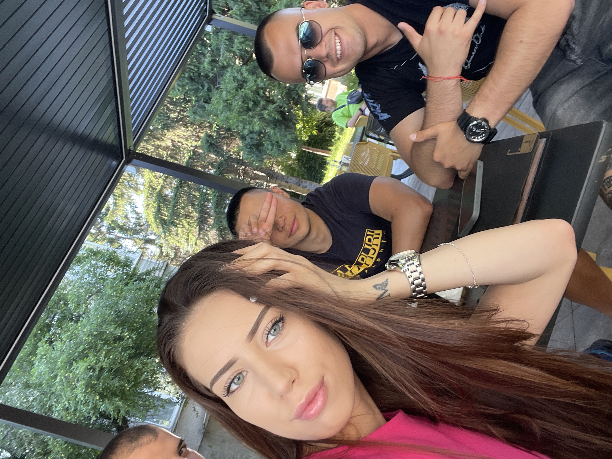 A selfie taken at an outdoor cafe shows four people. In the foreground, a woman with long brown hair and light blue eyes, wearing a bright pink top, smiles at the camera. Her left hand is resting on her forehead, and she has a watch and bracelet on her left wrist. To her right, a man wearing a black t-shirt and sunglasses smiles and gives a thumbs-up. His right hand is visible, also giving a thumbs-up. To the left of the woman, a man in a black t-shirt covers his eyes with his left hand. Behind them, another man in a bright green shirt sits at a table. The background consists of trees and a building. The setting appears to be a covered outdoor seating area.