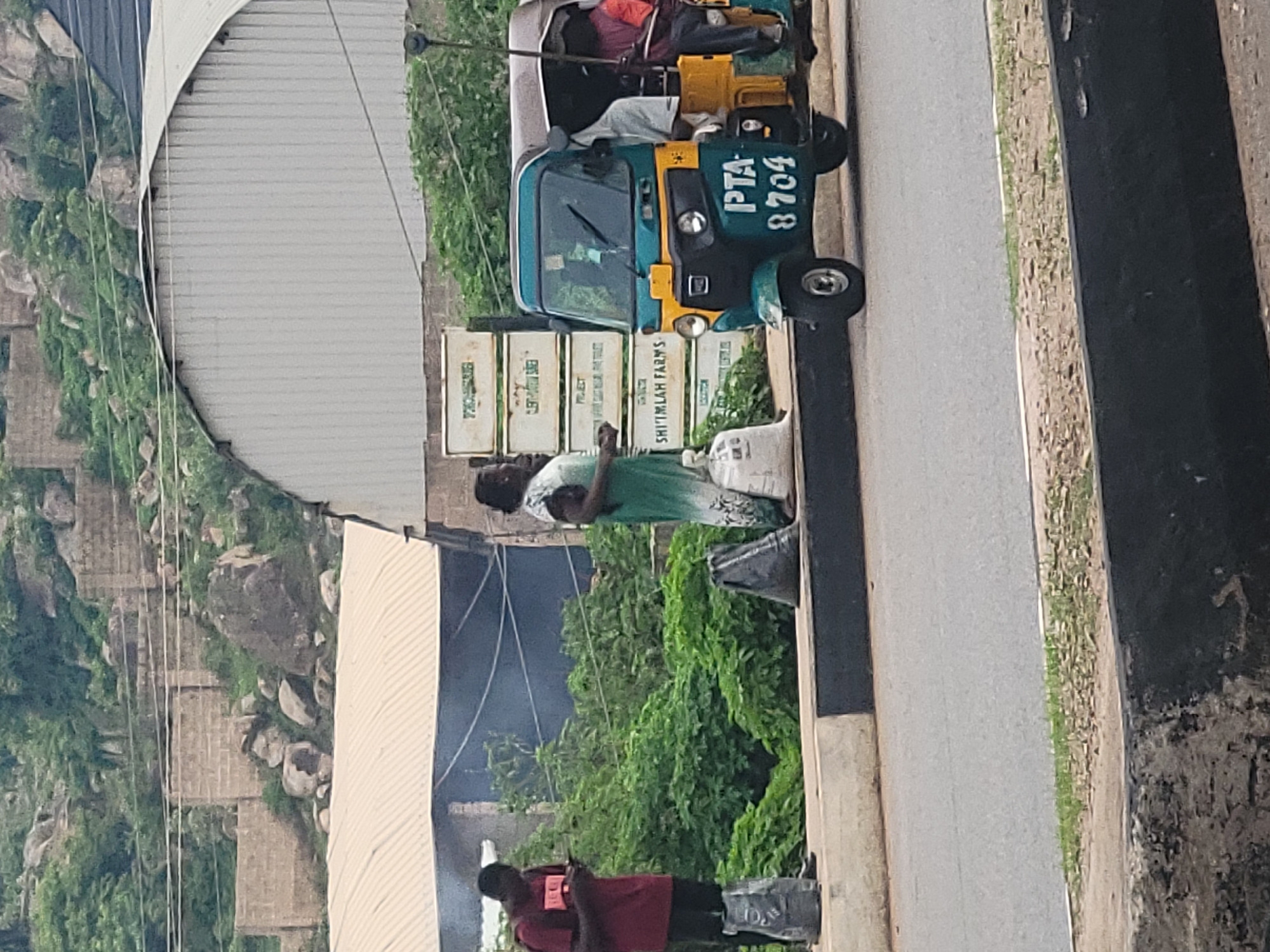 The image depicts a street scene in Gura Top, Nigeria. In the foreground, a paved road runs horizontally. To the left, a young man in a maroon t-shirt and black shorts stands on what appears to be two large bags, looking at his phone. Beside him is a black plastic bag. Further to the right, a woman in a light-colored, patterned dress stands next to several white crates stacked on top of each other. The crates have text visible, including "SHIMLAH FARMS." Adjacent to the crates is a green and yellow auto-rickshaw, also known as a Keke Napep, with the license plate "PTA 8704". Two people are visible inside the auto-rickshaw. The background shows lush green vegetation, some brick structures, and rocky hills. Smoke is visible rising from behind the green bushes and brick structures. The overall scene suggests a roadside waiting area or market stall.