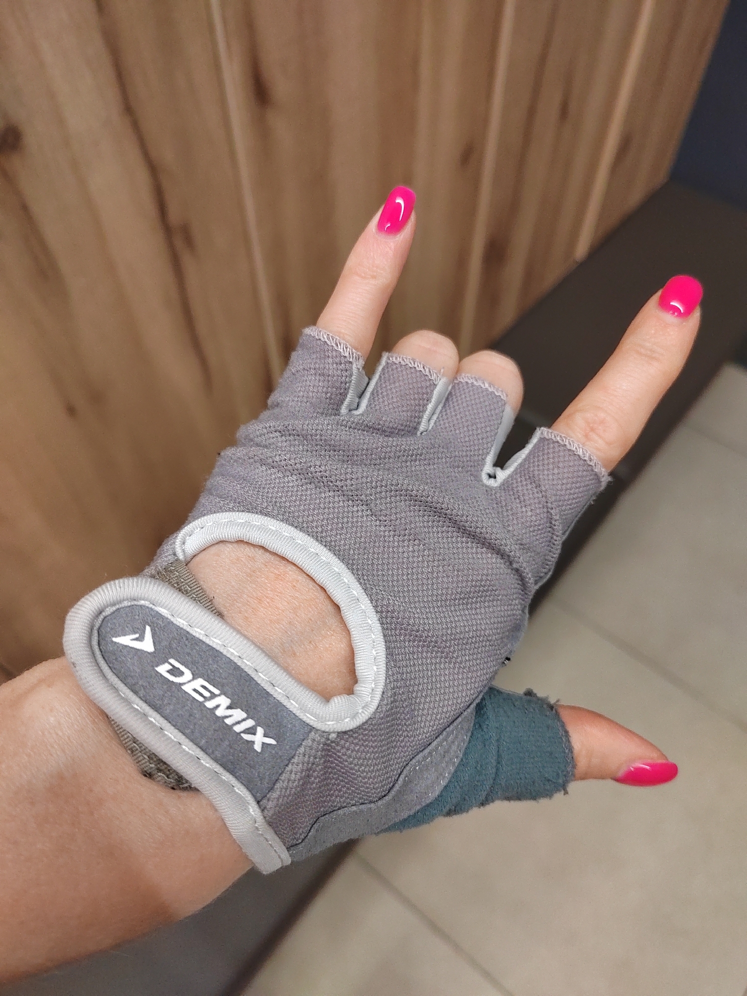 A hand wearing a gray fingerless glove with a white and gray logo that reads "DEMIX" is shown. The hand is making a rock and roll gesture, with the index and pinky fingers extended upwards. The fingernails are painted a bright pink color. The background consists of a wooden wall and a dark surface. The image appears to be a close-up shot. The context of the image's location within the city of Novogorelovo, Russia, is not determinable from the visual information provided.