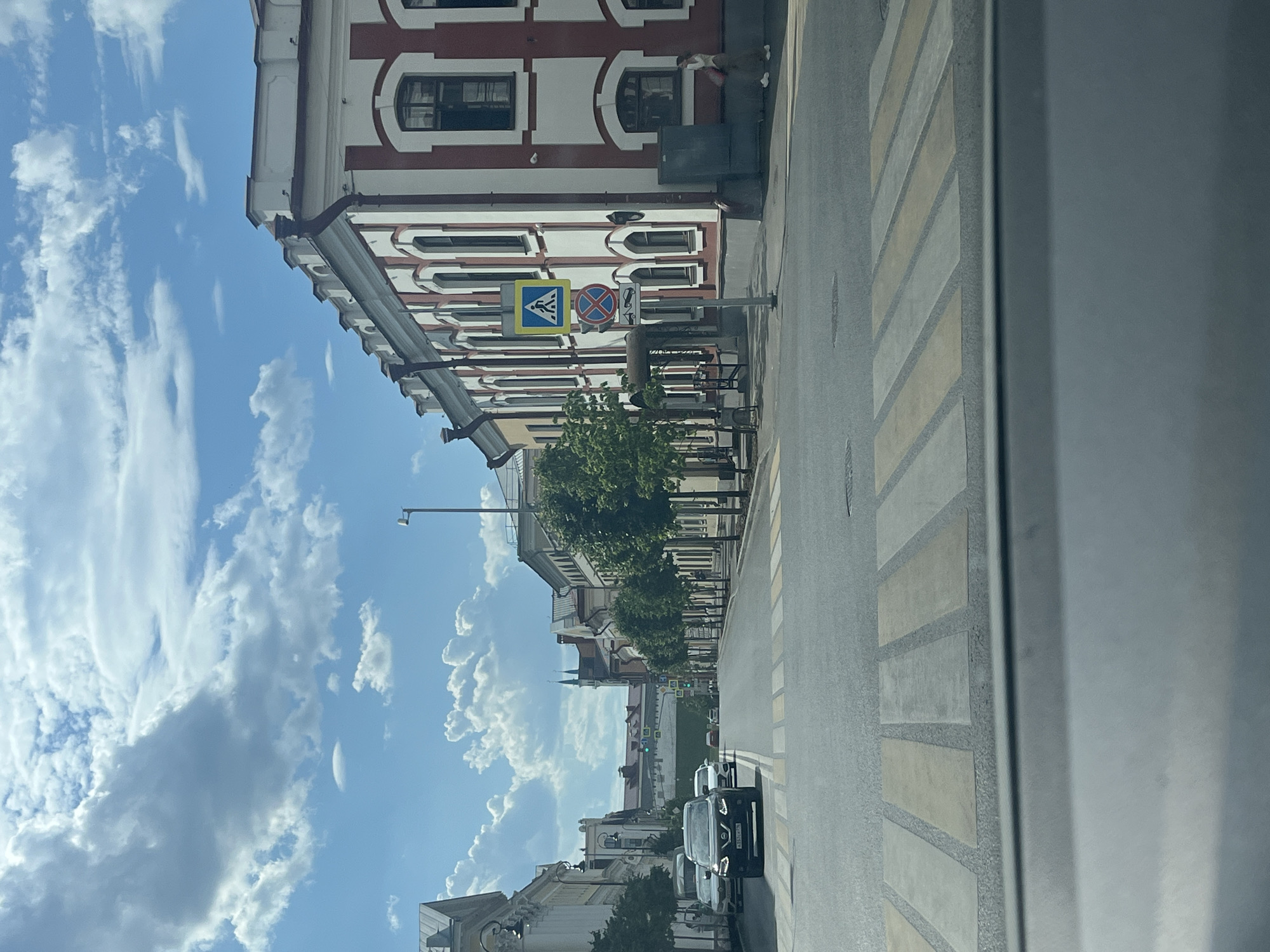 This media file depicts a street scene in Kazan, Russia. The primary subject is a street with a prominent pedestrian crossing. On the left side of the street, a building with red and white architectural detailing is visible, featuring numerous windows and decorative elements. Further down the street, more buildings line the road, interspersed with trees. On the right side of the street, the pedestrian crossing is marked with yellow and white stripes. A few cars are parked or driving on the street.
The sky is blue with a significant amount of white, fluffy clouds, suggesting it is daytime. The lighting indicates it is likely midday or afternoon. The scene appears to be set in an urban environment, possibly a historic district given the architecture of the buildings. There are no people clearly visible engaging in any activity, though the presence of a pedestrian crossing suggests people would be using the area.
Notable details include street signs. One sign is a blue square with a white pedestrian icon, indicating a pedestrian crossing. Another sign is a circular red sign with a white horizontal bar and a diagonal red line, likely indicating "no entry" or a similar prohibition. A license plate on a black SUV is partially visible, with Russian characters "A151CK716". The overall impression is of a bright, clear day in a city with traditional European-style architecture.
