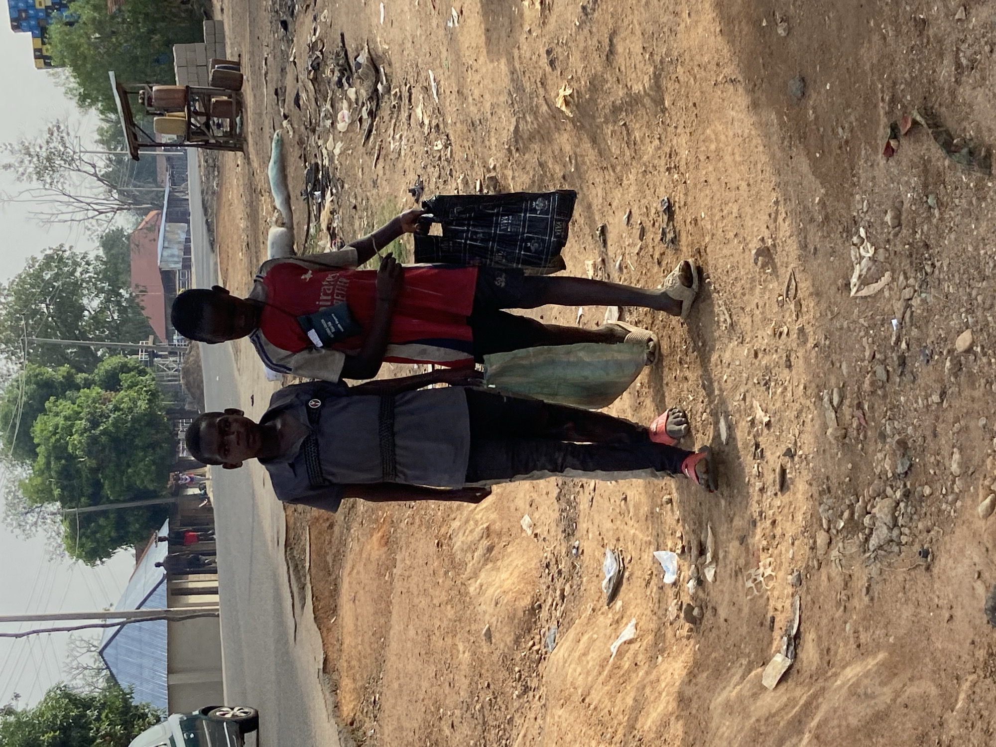 Two young males stand on a reddish-brown dirt path next to a paved road in Mararaba, Nigeria, under bright daylight.
The individual on the left (from the viewer's perspective if the image is rotated upright) wears a dark grey short-sleeved collared shirt with embellishments on the shoulders, dark trousers, and red sandals. He has a black strap around his waist and is carrying a large green container on his back, secured by shoulder straps, suggesting it might hold water or fuel.
The person on the right wears a red short-sleeved t-shirt with the text "emirates FLY BETTER" visible on the front, dark shorts, and light-colored sandals. He holds a black patterned plastic bag in his left hand and a smaller black pouch or bag in his right, which has the word "PAYA" repeated on it.
The immediate foreground and ground around them are unpaved, dusty, and littered with various small debris, papers, and rocks. In the background, to the left, a paved road is visible with a portion of a white car. Beyond the road, several buildings with corrugated metal roofs are nestled among green trees. Further down the dirt path, a rudimentary street stall is visible, featuring several yellow and brown jerry cans, commonly used for fuel or water. The overall setting appears to be a semi-urban or rural environment with signs of local commerce and daily life. The clear lighting indicates it is daytime, likely late morning or afternoon.