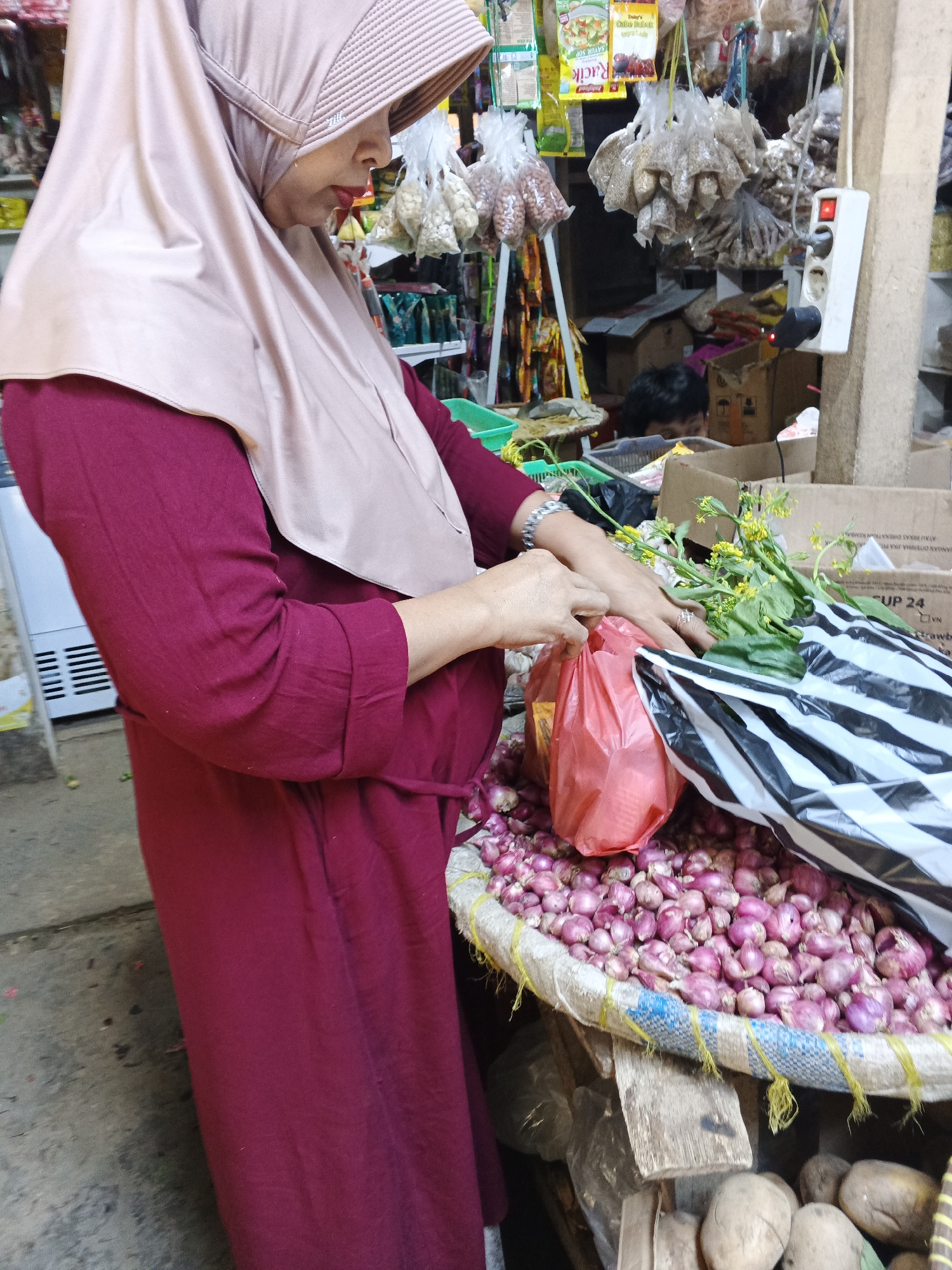 In Warungkiara, Indonesia, a woman wearing a maroon abaya and a beige hijab is standing in what appears to be a market stall. She is reaching into a black and white striped bag, placing items inside. In front of her, a large woven basket is overflowing with shallots. To her left, a red plastic bag sits on the shallots, and to her right, a bundle of green vegetables with small yellow flowers is partially visible. Behind the woman, various food items in plastic packaging are hanging, and in the background, there are cardboard boxes and a white electrical outlet with a red indicator light. The ground beneath the stall appears to be concrete.