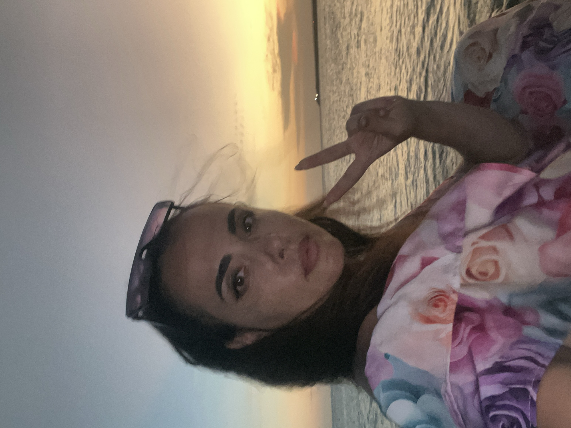 A woman is taking a selfie on a body of water at sunset. She is wearing a colorful, floral-patterned dress and sunglasses are resting on her head. Her right hand is raised, forming a peace sign. The sky is a gradient of blue and orange, and the water reflects the sunset colors. The scene appears to be a serene outdoor setting, possibly a beach or a boat. The background shows a distant shoreline with a few lights. The overall mood is relaxed and joyful. There is no indication of this location being in Kuta, Indonesia.