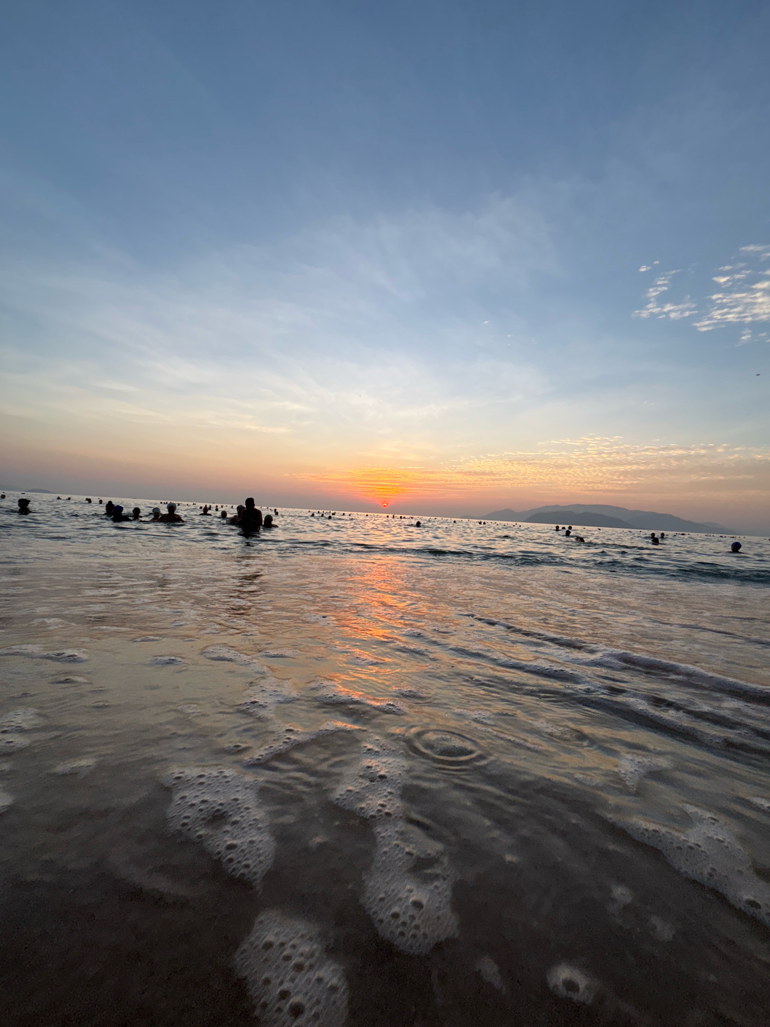 The image depicts a wide shot of the ocean at sunset. The sun is low on the horizon, casting a warm orange glow across the sky and reflecting on the water's surface. Several people are visible in the water, appearing to be swimming or wading. The foreground shows gentle waves lapping at the shore, with foam and bubbles visible. The overall atmosphere is serene and tranquil. This scene is consistent with the beaches of Nha Trang, Vietnam, known for their expansive coastlines and stunning sunsets.