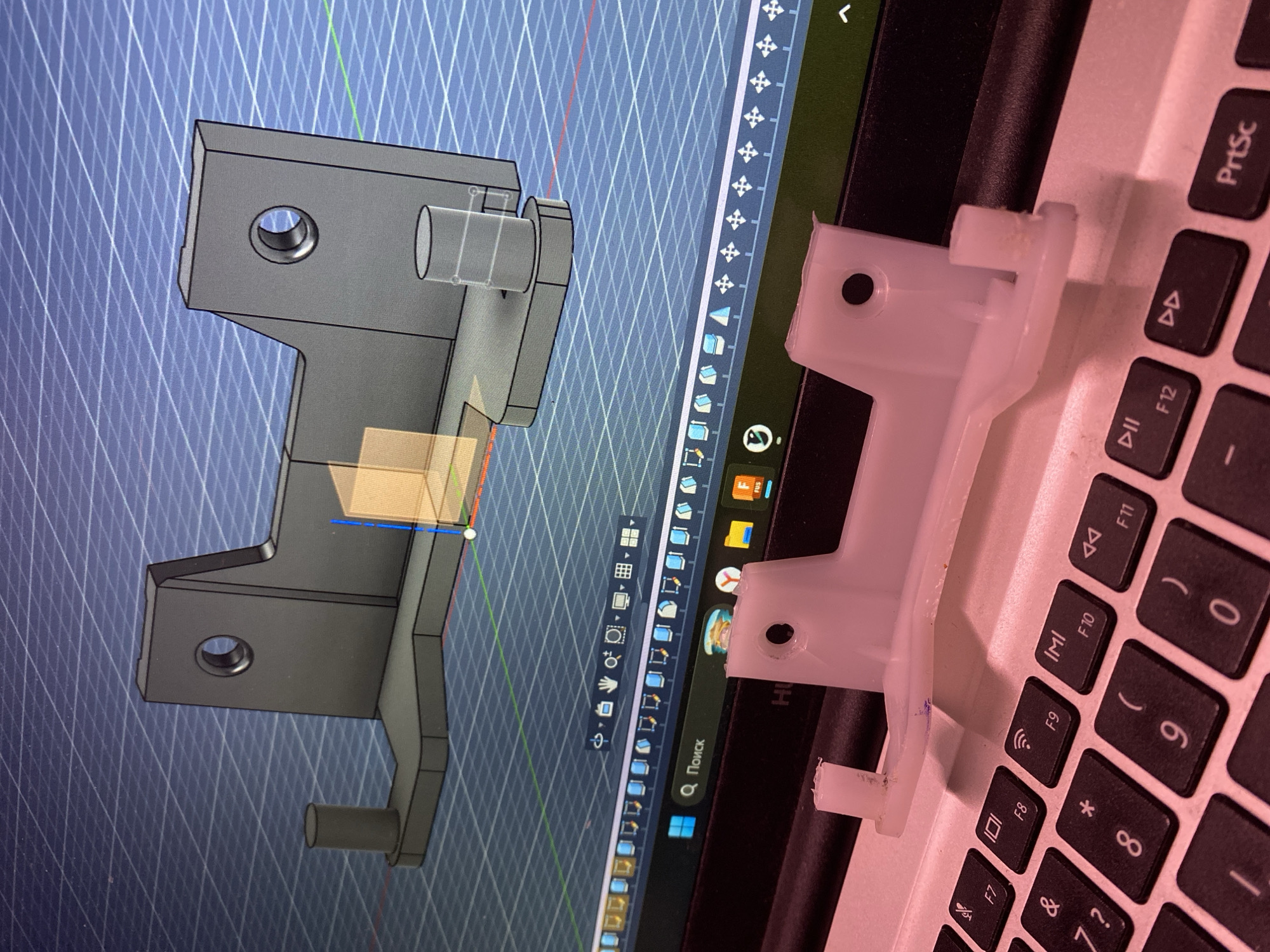A photograph captures a laptop screen displaying a CAD model of a bracket design on the left, while a 3D-printed version of the same bracket sits on the laptop's keyboard on the right. The CAD software presents a gridded background with the bracket rendered in grayscale, highlighting its various features, including cylindrical protrusions and mounting holes. Next to it, the physical bracket, likely made of white plastic, mirrors the design of the digital model, though it's slightly tilted on the keyboard, which includes keys such as "PrtSc," "F12," and "0." The laptop's bezel and the keyboard area appear black, and the screen also shows elements of the Windows operating system, including the search bar and various icons. The photo was taken in the city of Chaikovsky, Russia.