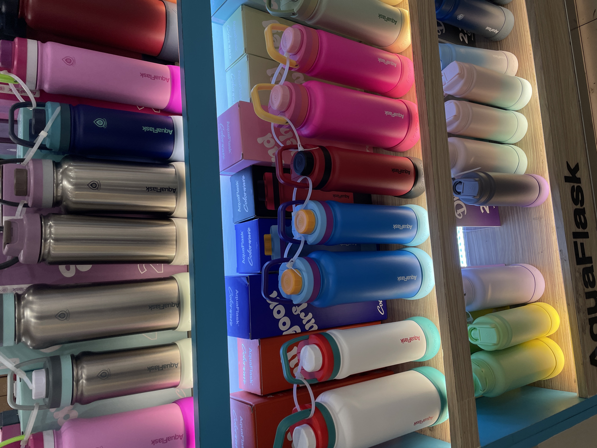 The image shows a display of AquaFlask brand water bottles on shelves, likely in a retail store. The bottles are of various colors, including pink, silver, blue, red, white, and pastel shades. They are arranged in rows on wooden shelves. Some bottles have handles, and others have caps with loops. The lighting suggests an indoor environment, possibly a mall or a specialty store. The text "AquaFlask" is visible on many of the bottles and on boxes in the background. The overall scene is a commercial display of merchandise. Without further context or visual cues in the image, it is not possible to determine the specific location within Davao City, Philippines.