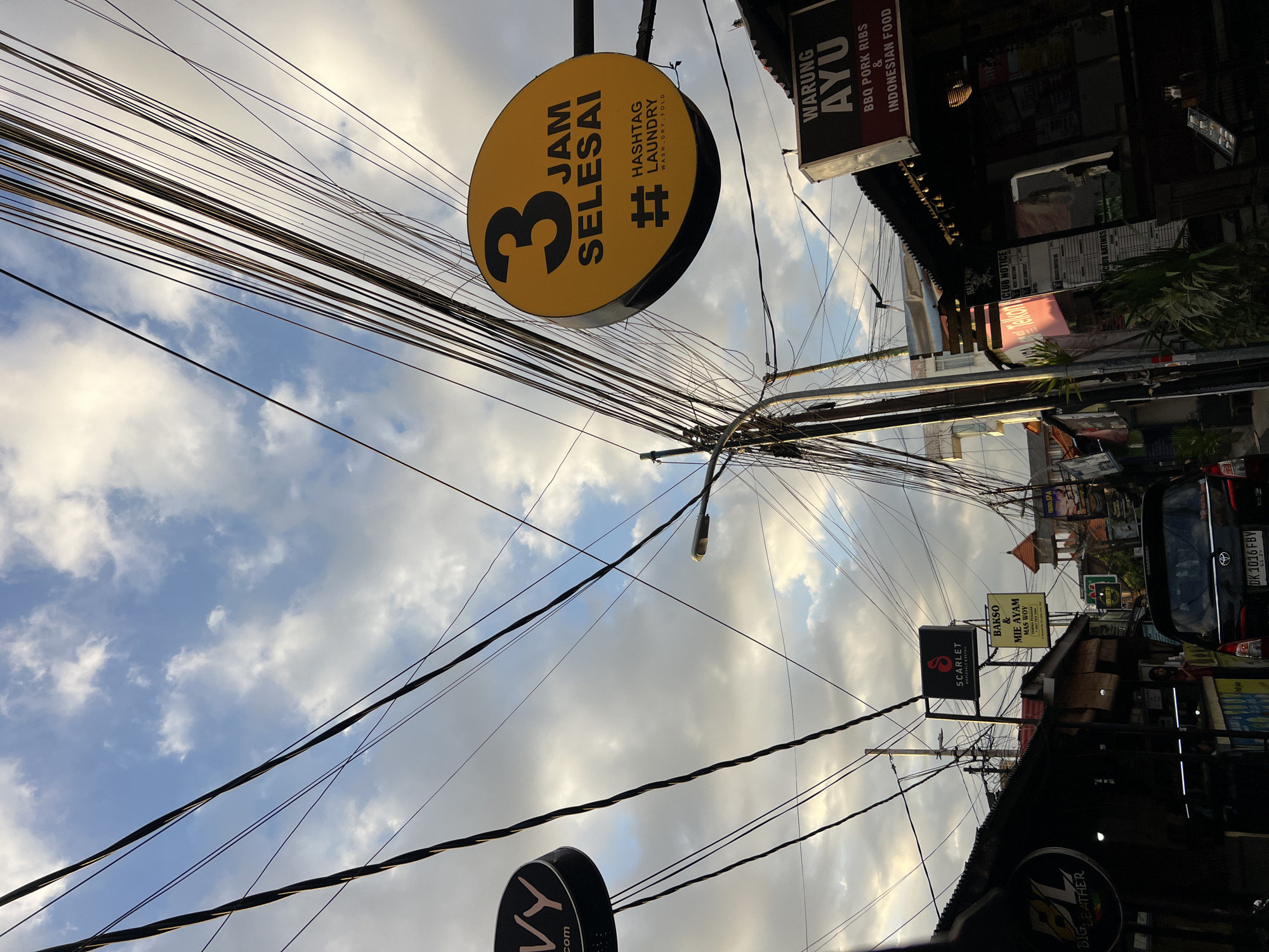 The image depicts a street scene in Kuta, Indonesia, characterized by a dense network of overhead electrical wires crisscrossing against a cloudy sky. The perspective is from street level, looking upwards. The street is lined with buildings, many featuring signs advertising various businesses. Prominent signage includes a yellow circular sign with "3 JAM SELESAI" and "# HASHTAG LAUNDRY WASH DRY FOLD," a black sign with "IVY.com," and a dark sign that reads "WARUNG AYU BBQ PORK RIBS & INDONESIAN FOOD." Other visible signs include "SCARLET WHOLESALE & RETAIL" and a sign for "BIG LEATHER." A black car is visible in the lower center of the frame. The overall impression is of a busy urban environment with the infrastructure of electricity and commerce prominently displayed.