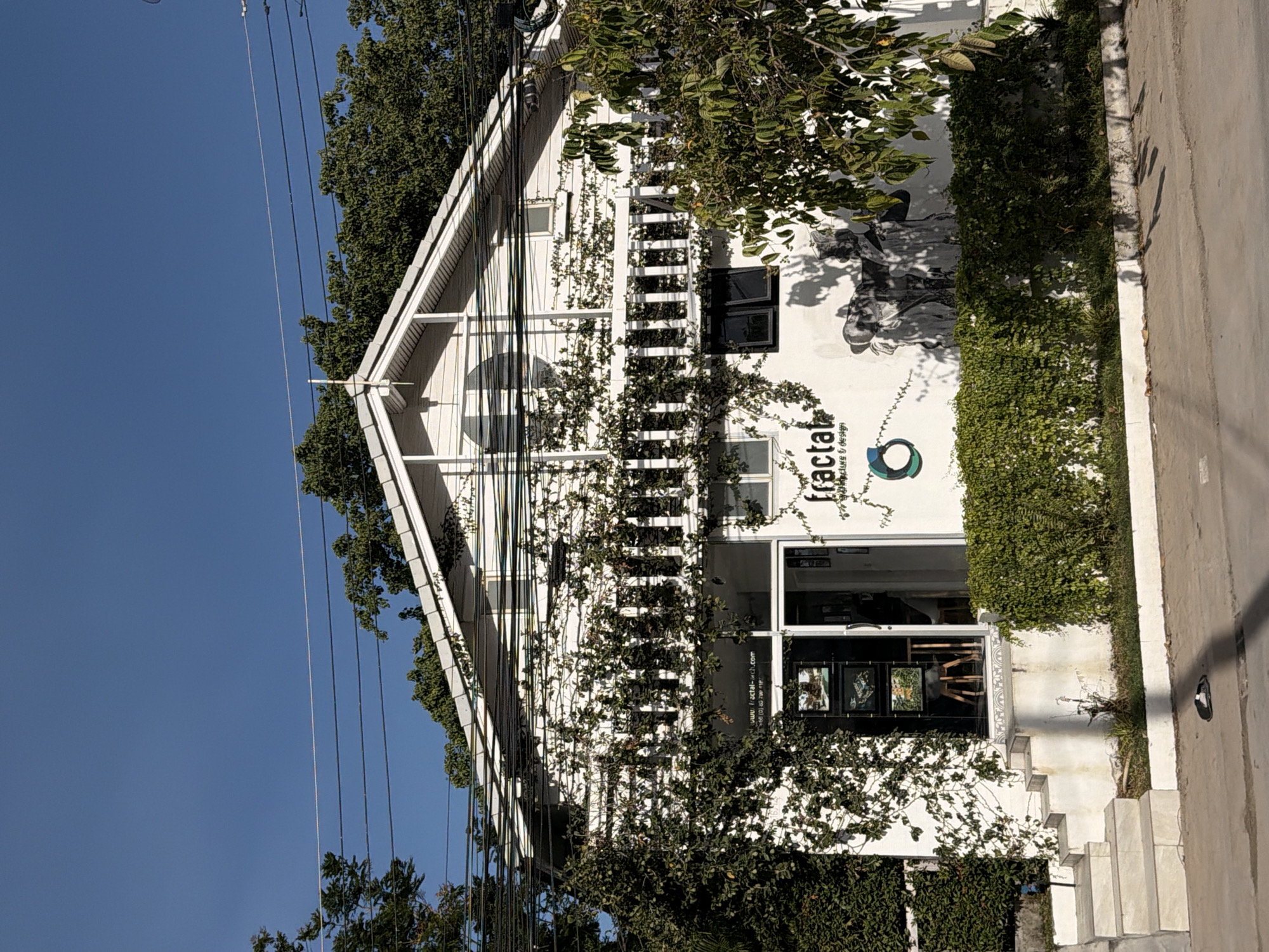 The image shows the exterior of a white, two-story building, which appears to be a business or gallery, located in the city of Ko Samui District, Thailand. The building has a peaked roof and is partially covered in green vines. There are several windows and a glass-paneled door on the ground floor. A white railing with vertical slats runs across the second floor. Text on the building reads "fractal architecture & design". A mural of what appears to be two figures is painted on the white wall to the right of the entrance. In front of the building is a low hedge, and to the left, there are stone steps leading up to the entrance. The ground in front of the building is paved, and a black and white object, possibly a sandal, is lying on the pavement. Overhead, there are multiple electrical wires strung across the scene. The sky is clear and blue. Trees surround the building and vegetation is abundant.