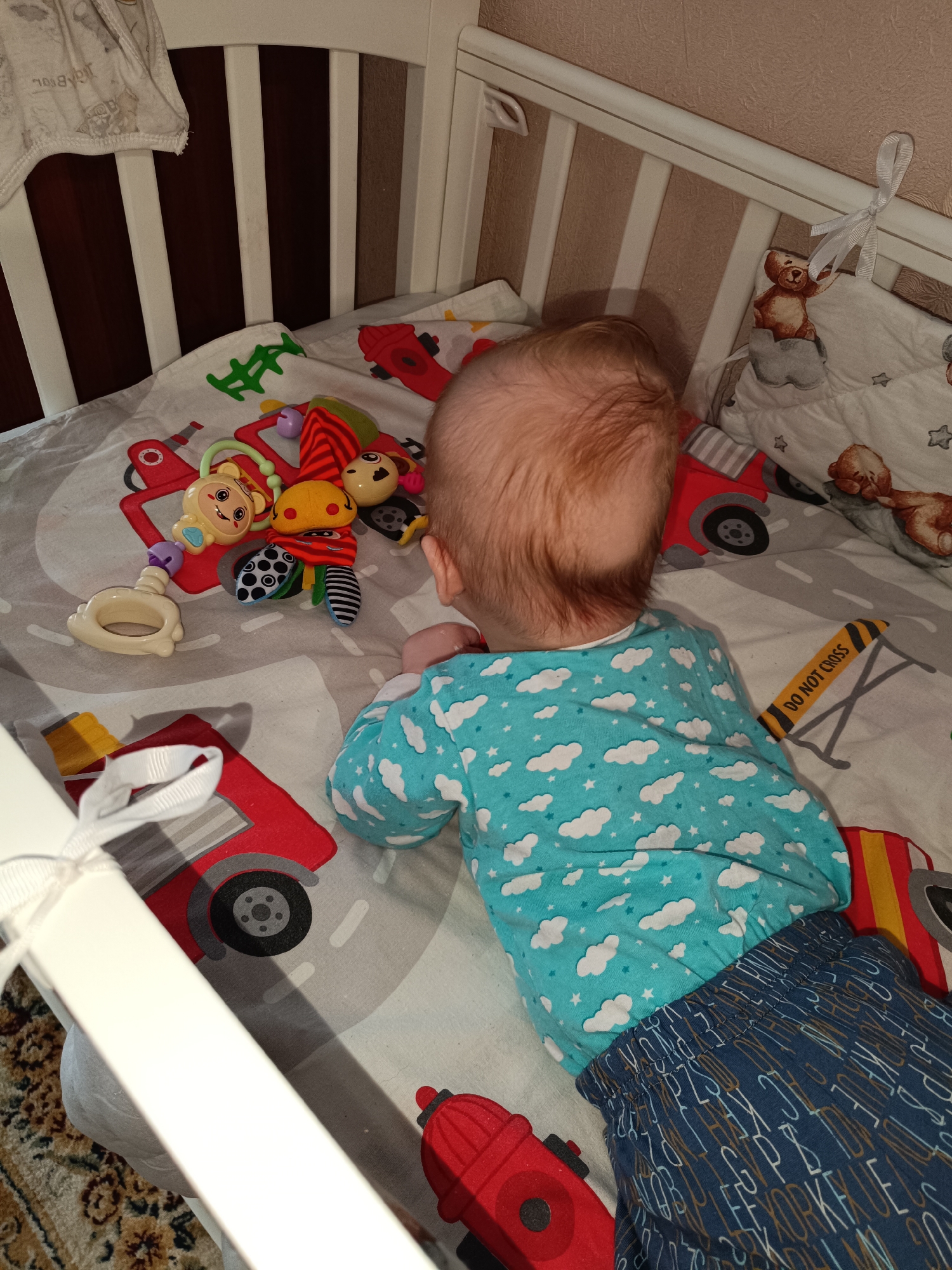 A baby is lying on their stomach in a crib. The baby is wearing a blue pajama top with white cloud and star patterns and dark blue pants with white and brown letter patterns. The crib has white railings and is lined with a sheet that has a road pattern and images of red firetrucks and other transportation-related graphics. There are baby toys scattered around the baby on the sheet. The location is likely a private residence or nursery in Tobolsk, Russia.