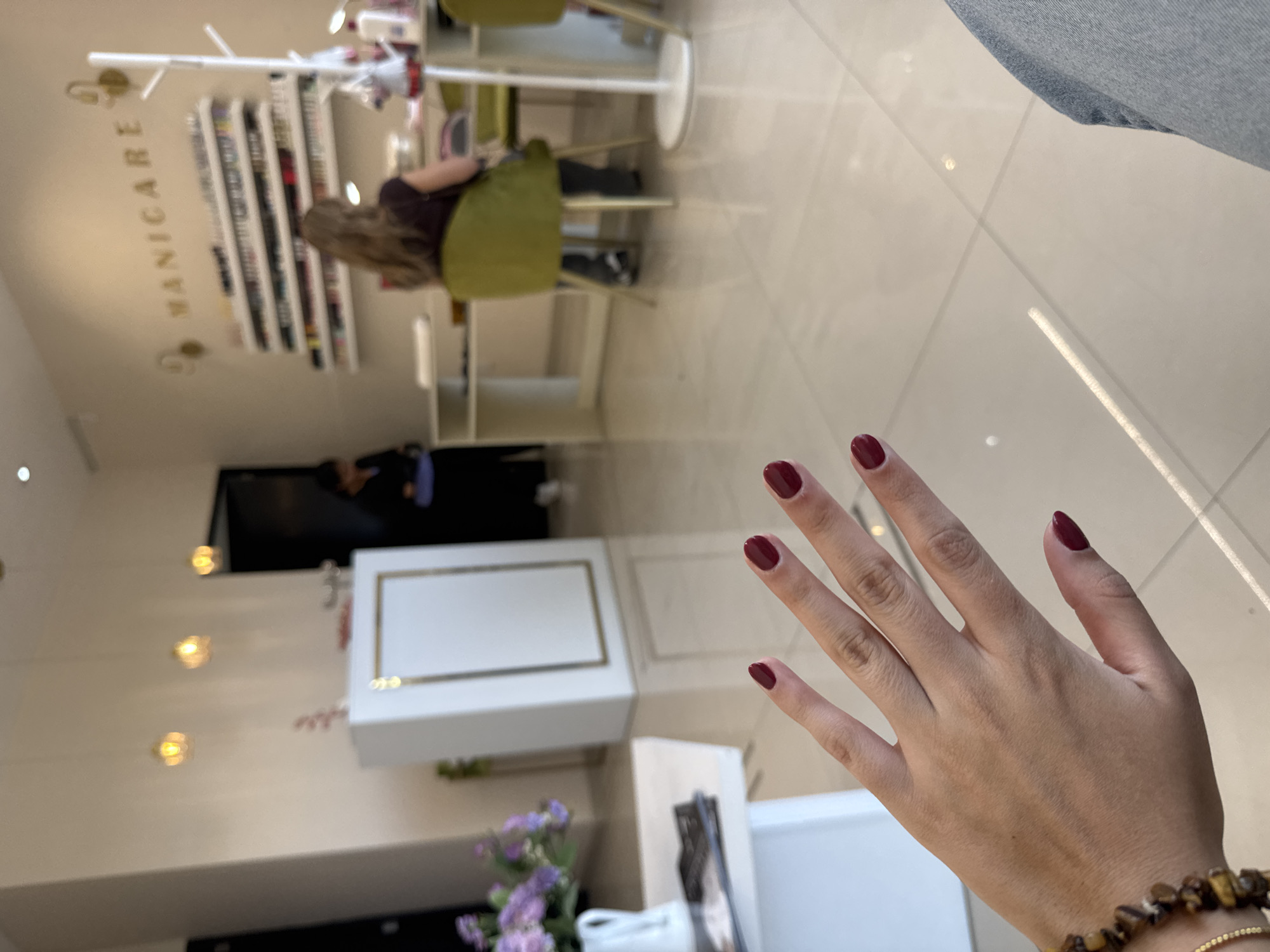A woman is in a nail salon. She is displaying her hand with newly manicured nails, painted a deep red color. In the background, another woman is sitting in a chair. A wall display of nail polish colors is visible, and the word "MANICARE" is on the wall. The location appears to be an indoor setting, presumably a nail salon in San Bernardino Tlaxcalancingo, Mexico.
