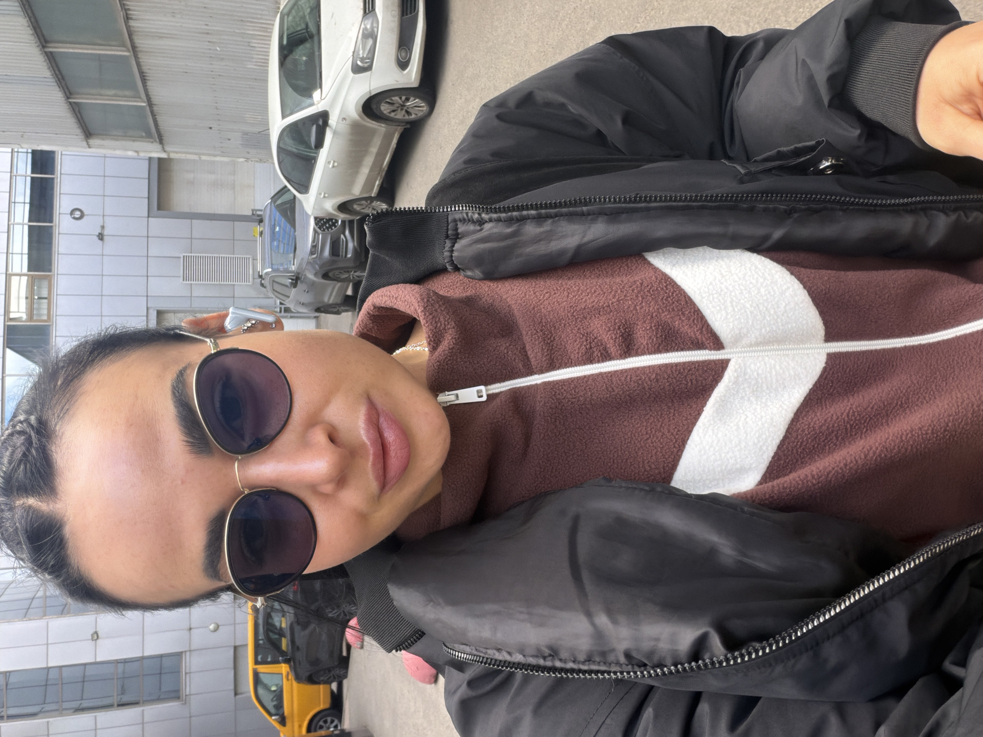 A person wearing sunglasses, a brown fleece jacket with a white stripe, and a black jacket is in an urban environment that might be in Saint Petersburg, Russia. A white car, a grey car, and a yellow car are nearby. The person is in front of a building with a series of vertically oriented windows.