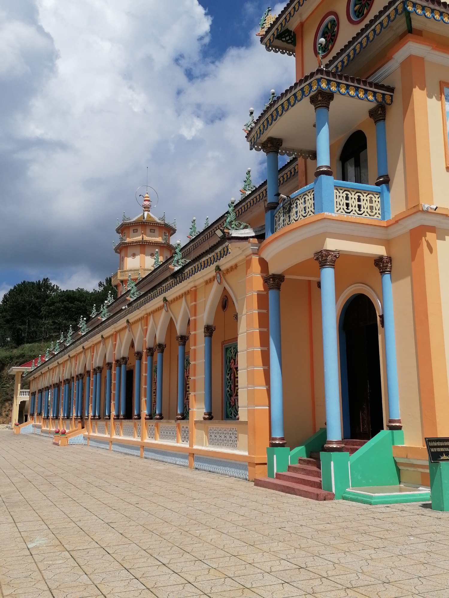 The image captures the exterior of a vibrantly colored building, likely a temple or pagoda, in Xuân Thành, Vietnam. The architecture is characterized by ornate detailing, with prominent blue and orange hues dominating the structure. A long colonnade of blue columns, topped with golden decorative elements, runs along the side of the building, supporting an orange roof with intricate patterns. Above the colonnade, a balcony features a blue railing with decorative cutouts. The building has a distinct pagoda tower in the background, also featuring the characteristic yellow and orange color scheme. The foreground is paved with light brown square tiles, creating a walkway. Lush green trees are visible on a hillside behind the building. The sky is a mix of blue with fluffy white clouds. A sign with text is visible on the right side of the image. There are no people or animals present in the scene.