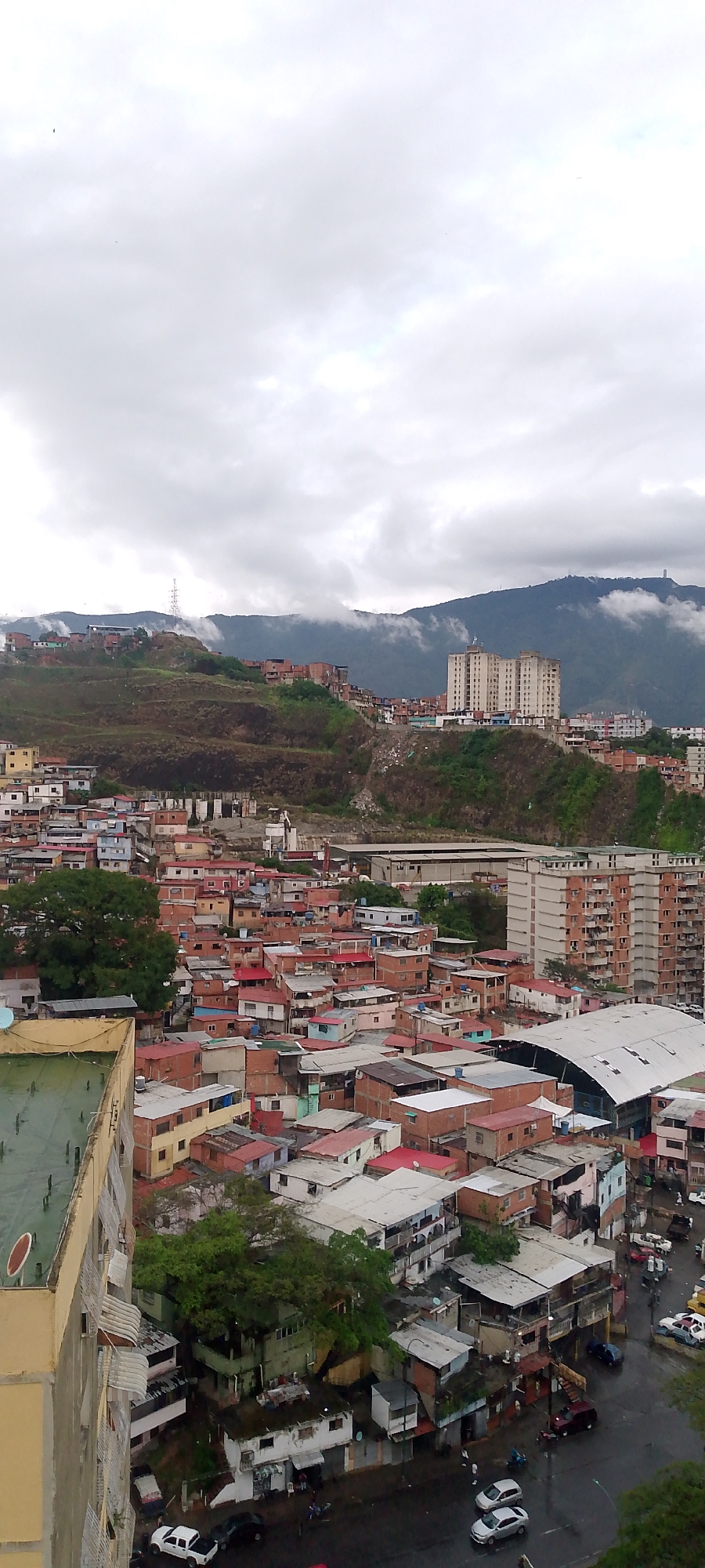 This is an aerial photograph of a densely populated hillside neighborhood in Caracas, Venezuela. The scene depicts a sprawling urban environment with numerous small, red-tiled roof houses packed closely together, many of them constructed with exposed brick. These structures climb the side of a steep, barren hill. In the midground, a large, modern apartment building stands out, contrasting with the informal settlements. Further up the hill, the landscape is dominated by green mountains partially obscured by low-lying clouds. In the foreground, a portion of a yellow building with a green roof is visible, suggesting a higher vantage point for the photograph. Several cars are parked on a wet street at the bottom of the scene, indicating it may have recently rained. The sky is overcast with dense grey clouds.