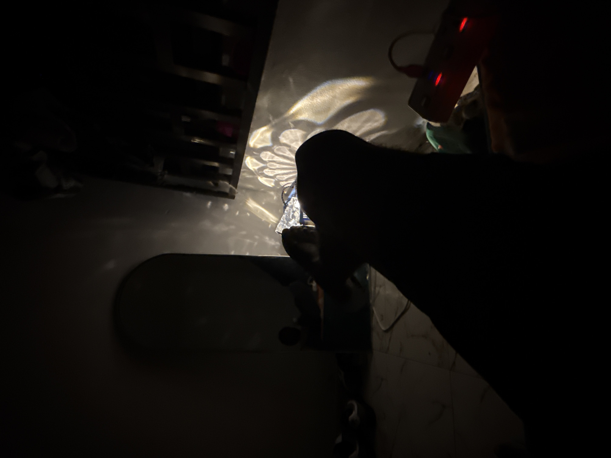 A silhouette of a dark-colored dog is visible in the foreground, facing away from the camera. The dog's head is turned to the right, and its ears are erect. In the background, there is a projected light pattern on a wall, resembling a flower or a sunburst. To the right of the projected light, a red power strip with illuminated red lights is partially visible. The environment appears to be a dimly lit room, possibly a bedroom, with a tiled floor. A large, dark, arched object, possibly a mirror or a wardrobe, is positioned on the left side of the frame. There are also some indistinct objects on the floor, including what appear to be shoes. The overall scene is dark, with the primary light source originating from a device casting the patterned light on the wall. The location is identified as Uyo, Nigeria.