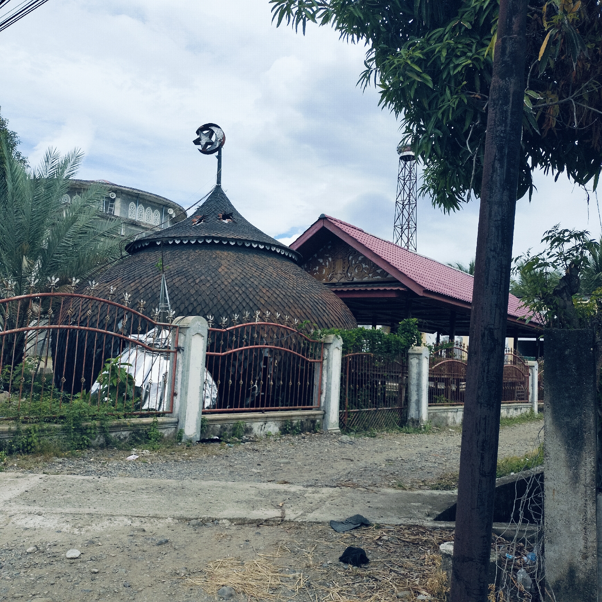 The image captures a static outdoor scene featuring a mosque complex in Kuta Pungwa, Indonesia, under a cloudy sky. The primary subject is a mosque building with a distinctive, large, dark-tiled, dome-like roof, possibly made of shingles or scales, crowned with a weathered metal crescent moon and star finial. A decorative fringe runs along the base of this dome.
In front of the mosque is a rusty metal fence with pointed decorative elements, set between concrete posts. Behind this fence, some greenery and shrubs are visible. To the right of the main dome, another building with a steep, red-tiled gabled roof is partially seen, featuring intricate wooden carvings under its eaves. Further back and to the right, a tall, slender metal lattice minaret extends into the sky.
On the left side, lush green palm fronds and other tropical tree leaves obscure part of another rounded building with multiple arched windows adorned with white, patterned grilles. A thick, dark utility pole stands prominently on the right side of the foreground, partially obscuring the view and rising towards other power lines crossing the top left of the frame.
The ground in the foreground consists of a rough dirt and gravel path or road, with patches of concrete and scattered debris like straw and dark fabric. No human activity or visible text is present in the image. The diffused lighting suggests an overcast daytime setting.